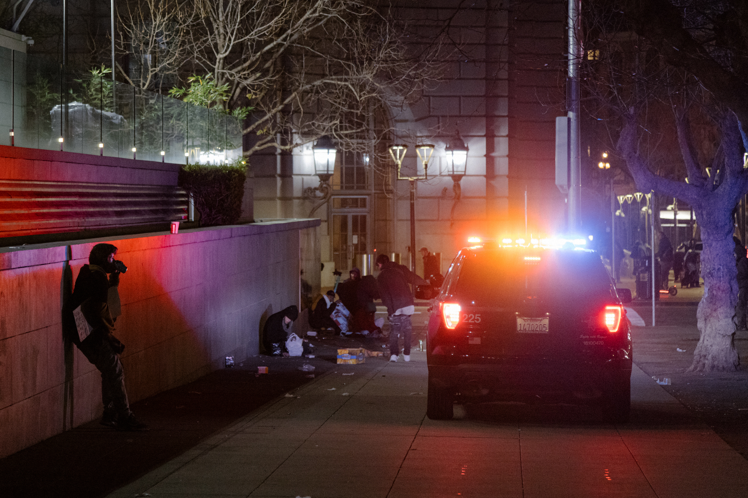 A police car with flashing lights is parked on a dimly lit street beside a building. People are gathered on the sidewalk, and a person is leaning against a wall.