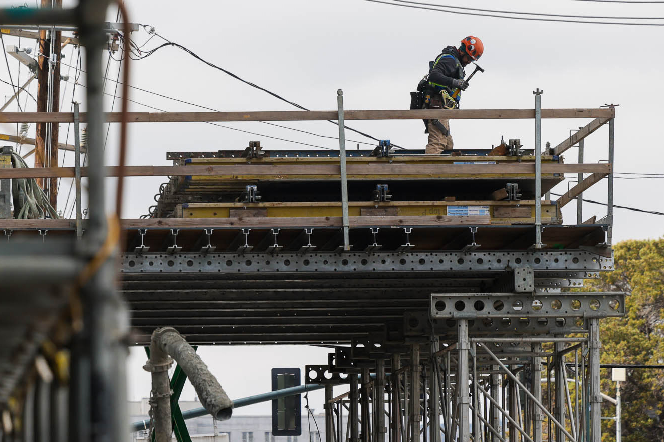 A construction worker in an orange helmet and safety gear stands on a scaffold, surrounded by tools and equipment, with power lines and trees in the background.