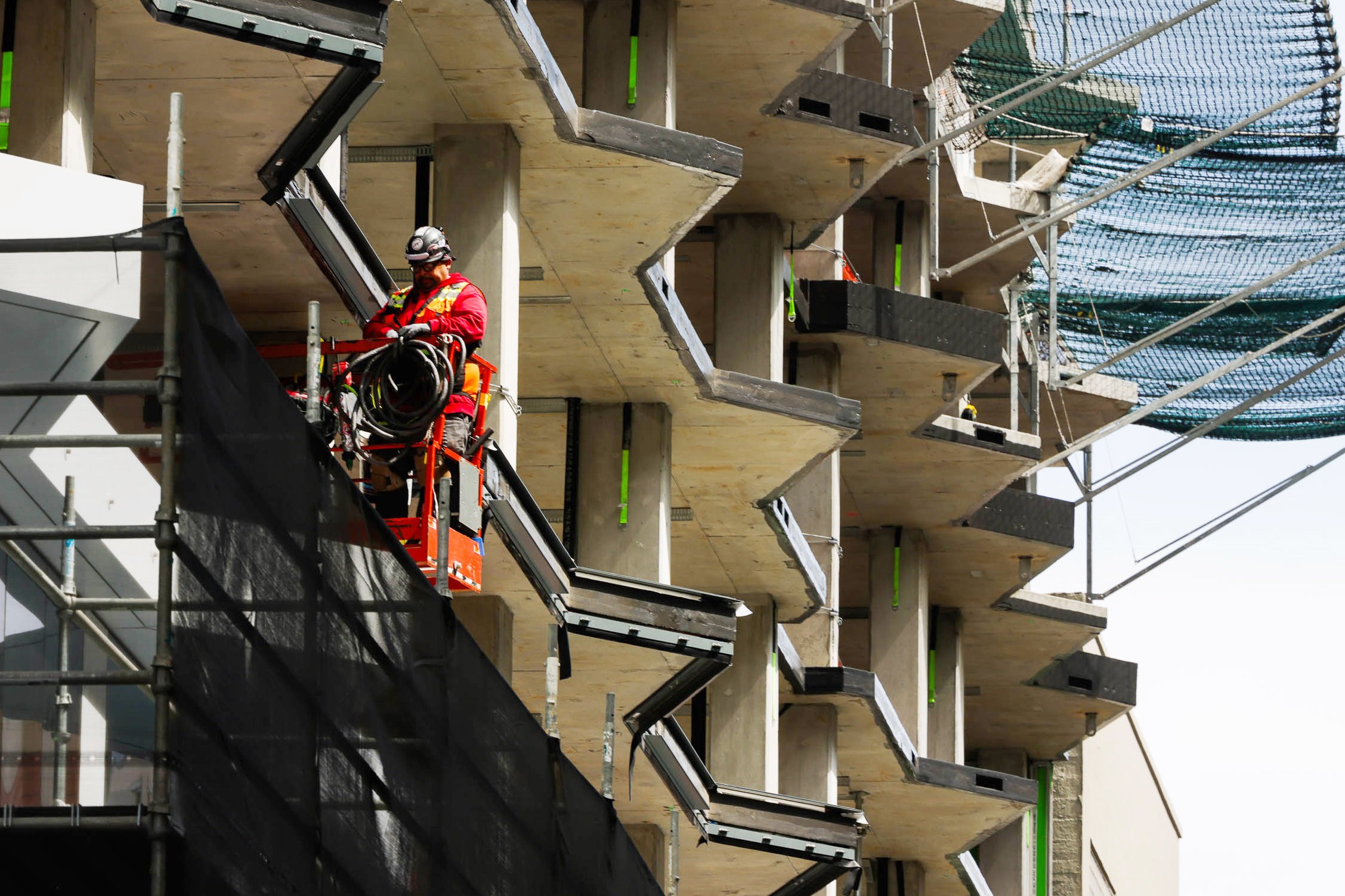 A construction worker in red outfit and safety gear operates a lift near a modern building with staggered concrete balconies and scaffolding with netting.