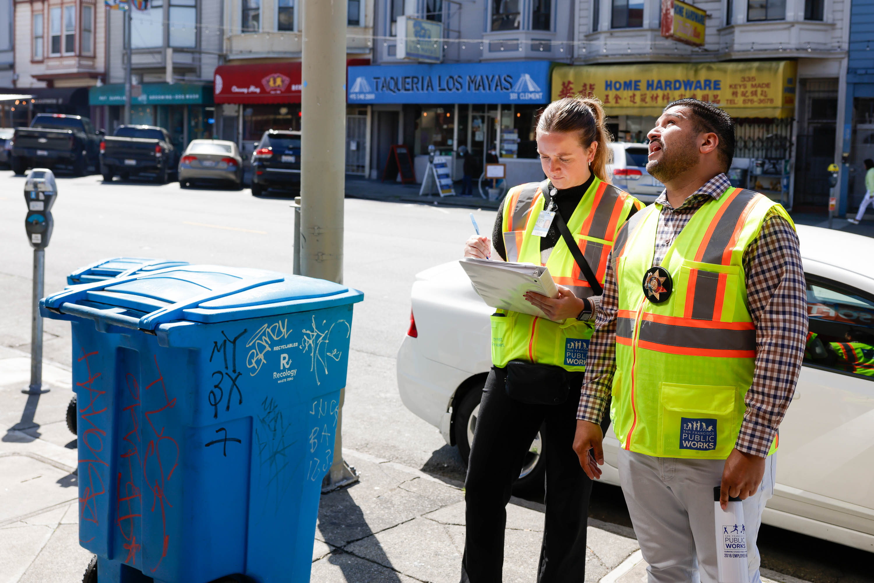 Two people in safety vests inspecting a blue recycling bin tagged with graffiti on a sunny street.
