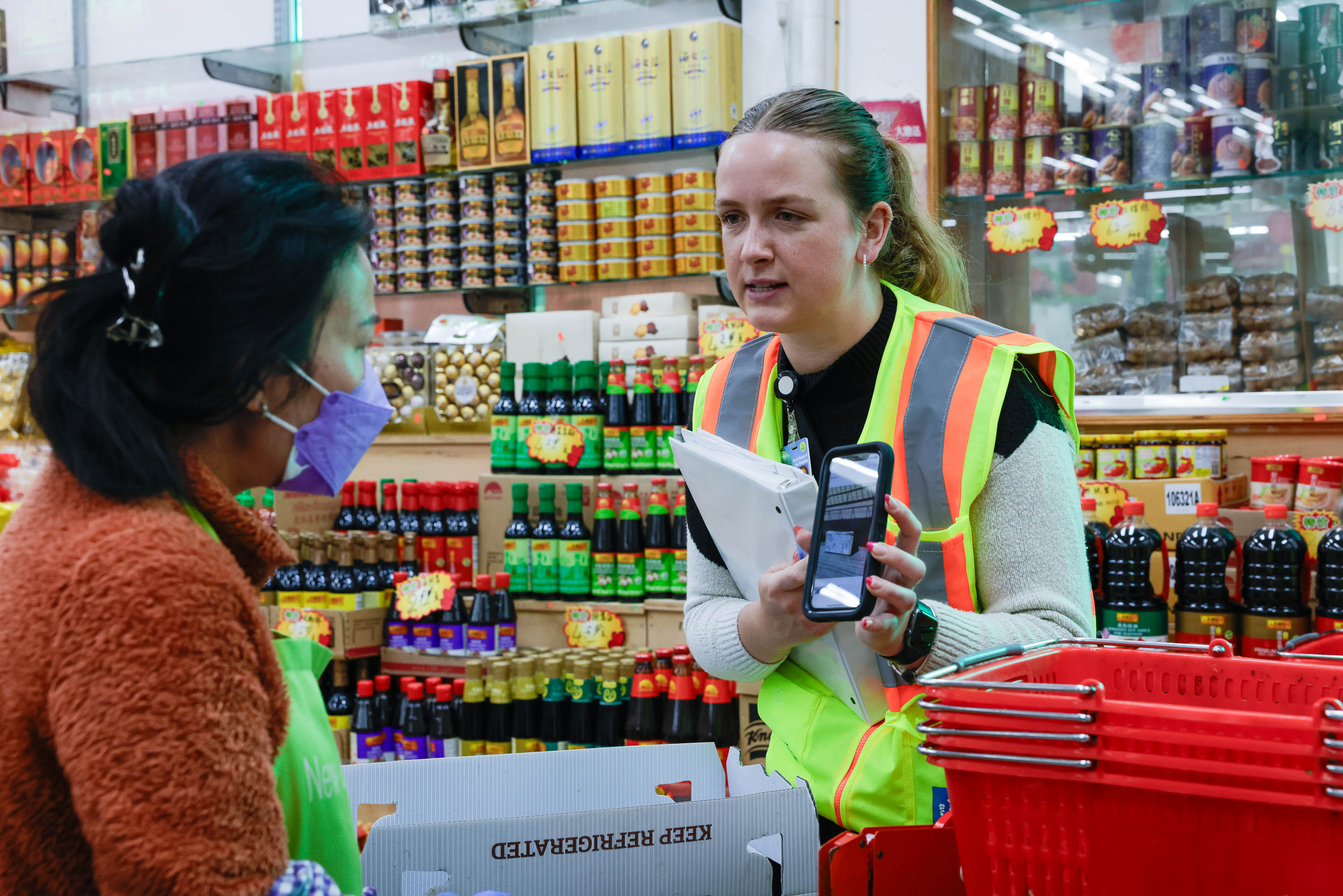 Two women converse in a store, one in a high-visibility vest holding a phone; shelves with goods surround them.