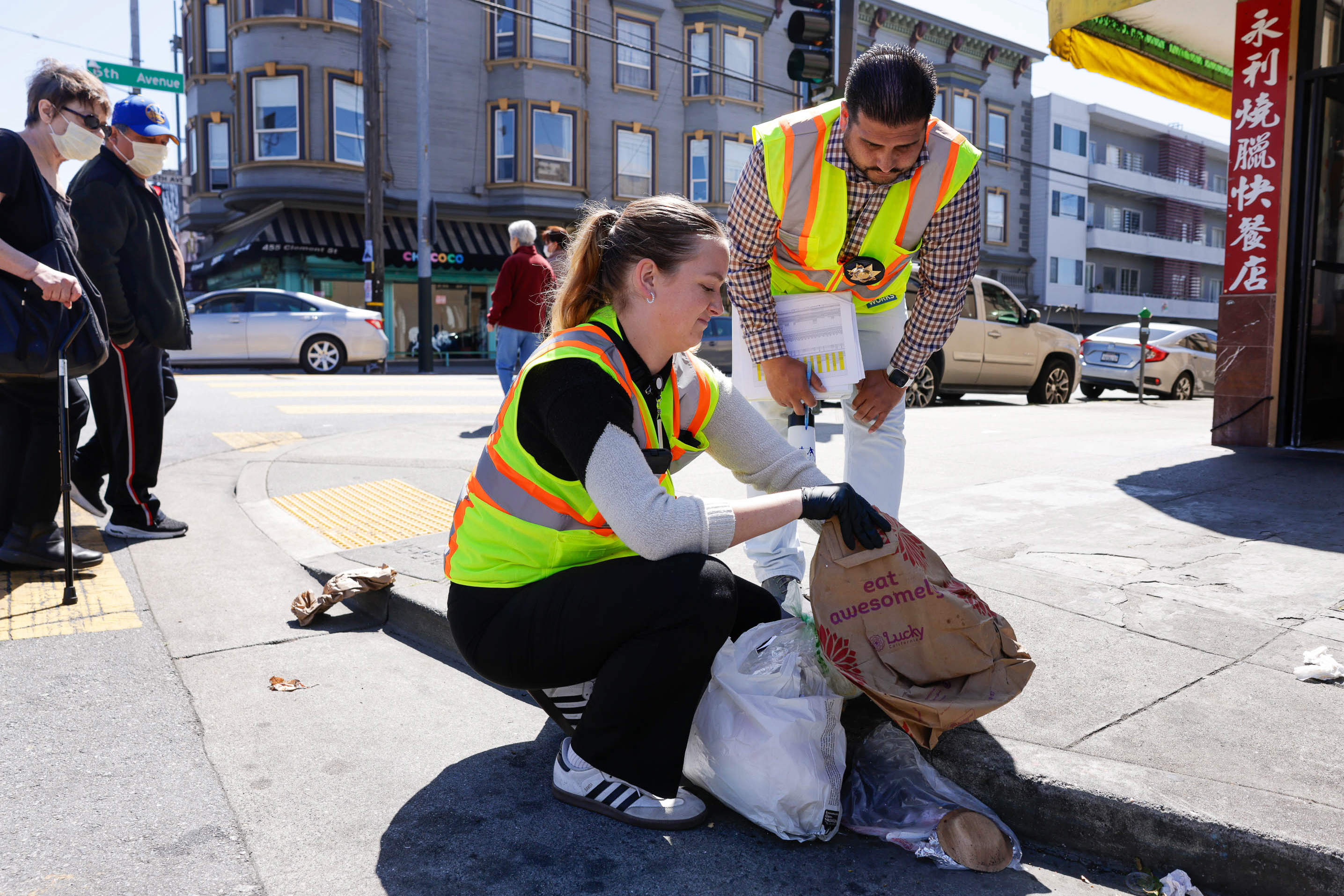 Two volunteers in reflective vests clean a city street corner, collecting litter into bags.