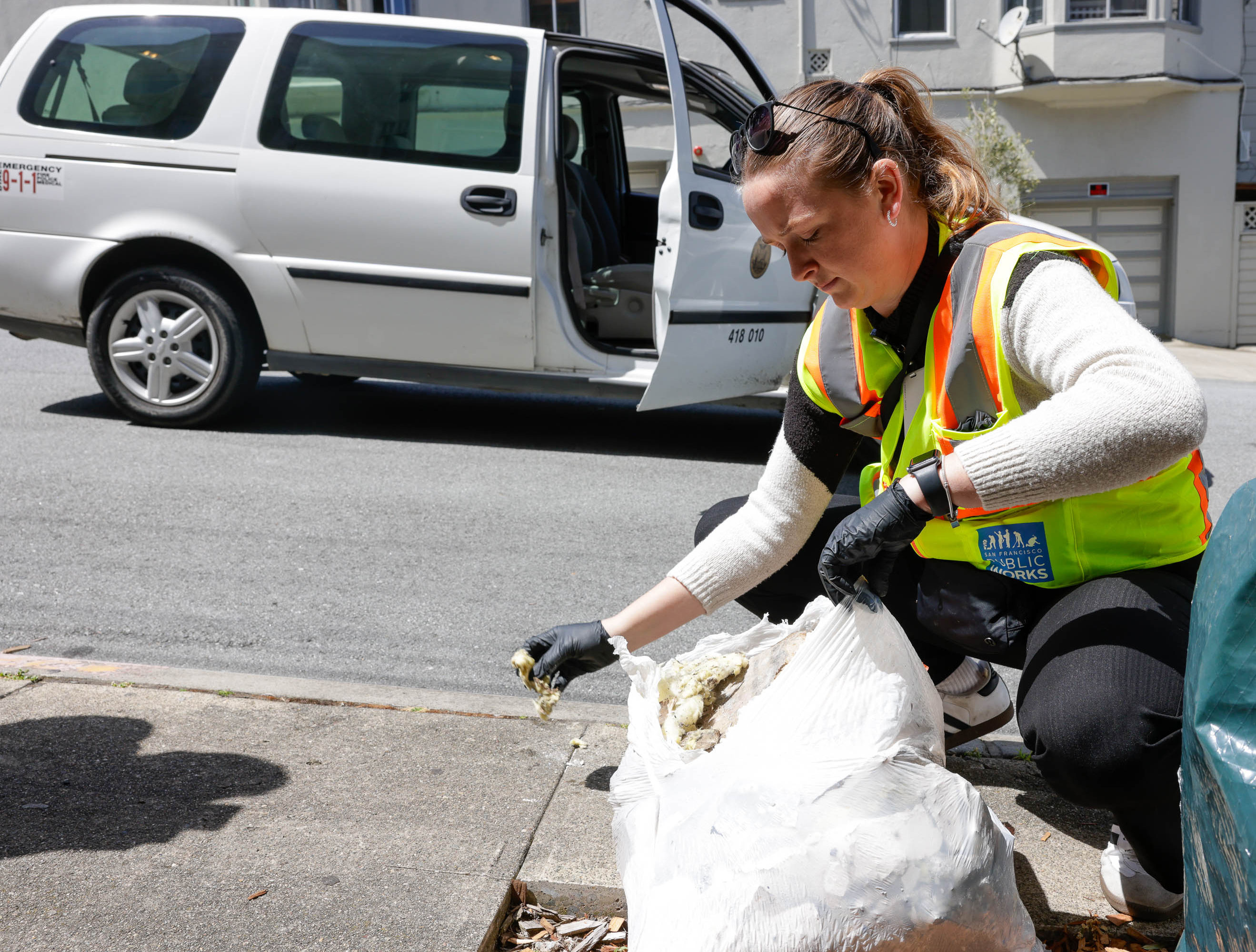 A woman in a safety vest cleans street debris near a parked van.