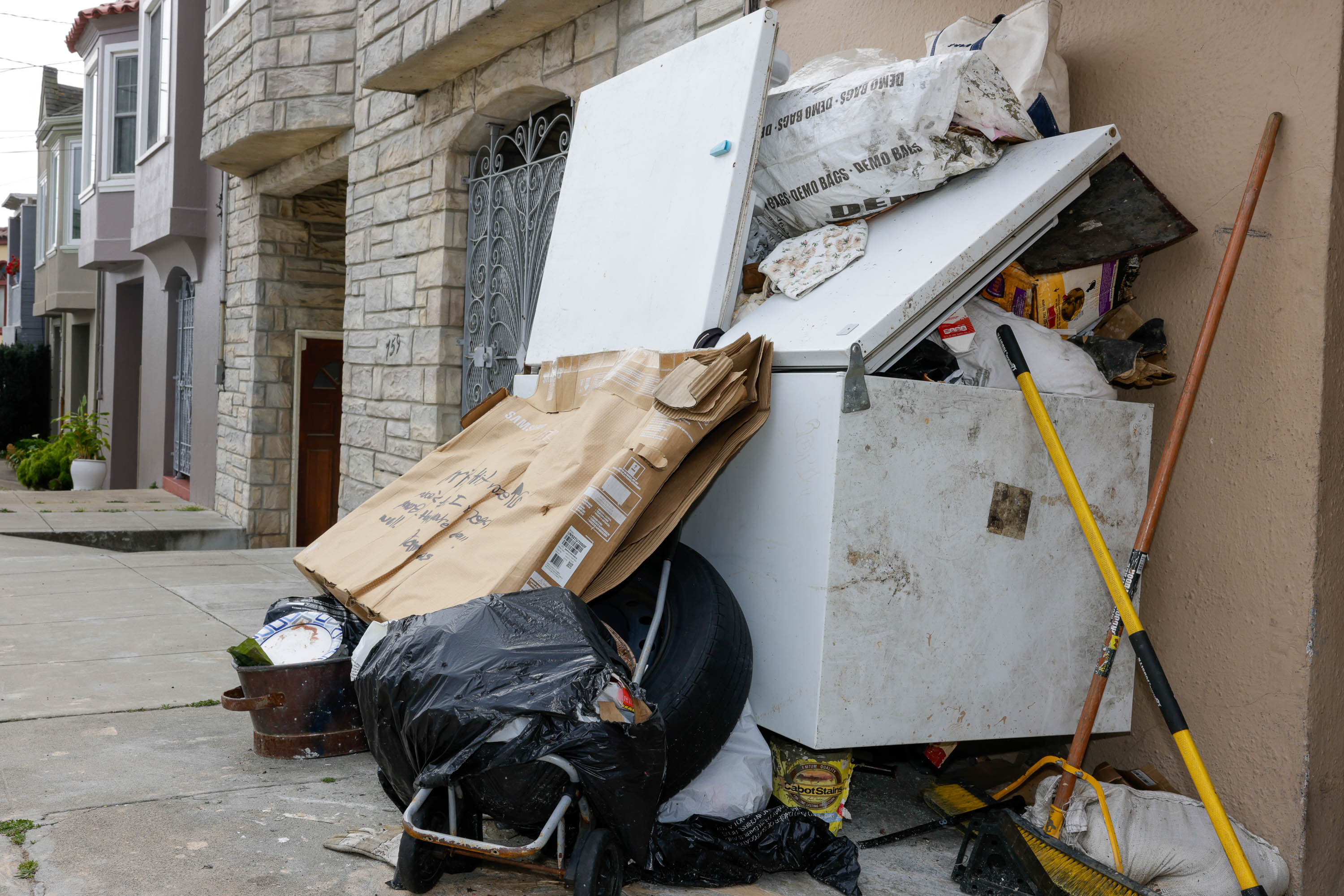 A cluttered sidewalk with a fridge, garbage bags, cardboard, and construction tools.