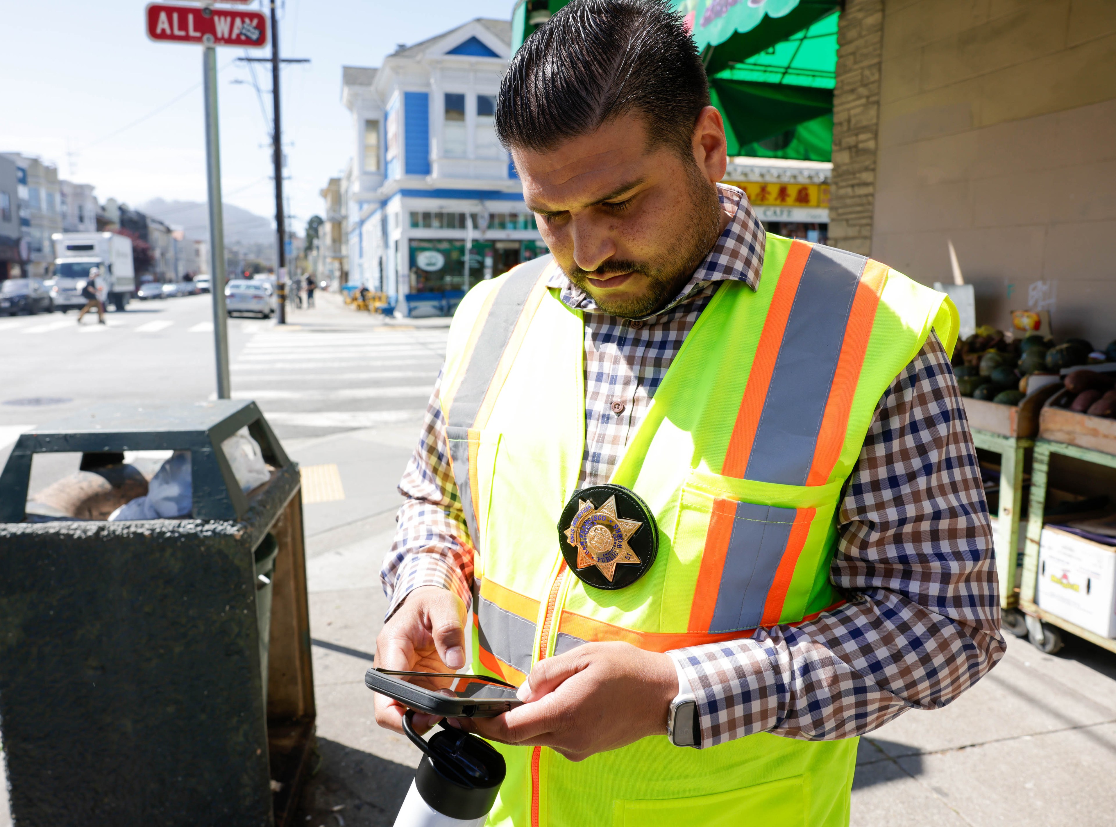 A man in a high-vis vest with a badge looks at his phone on a sunny street corner.