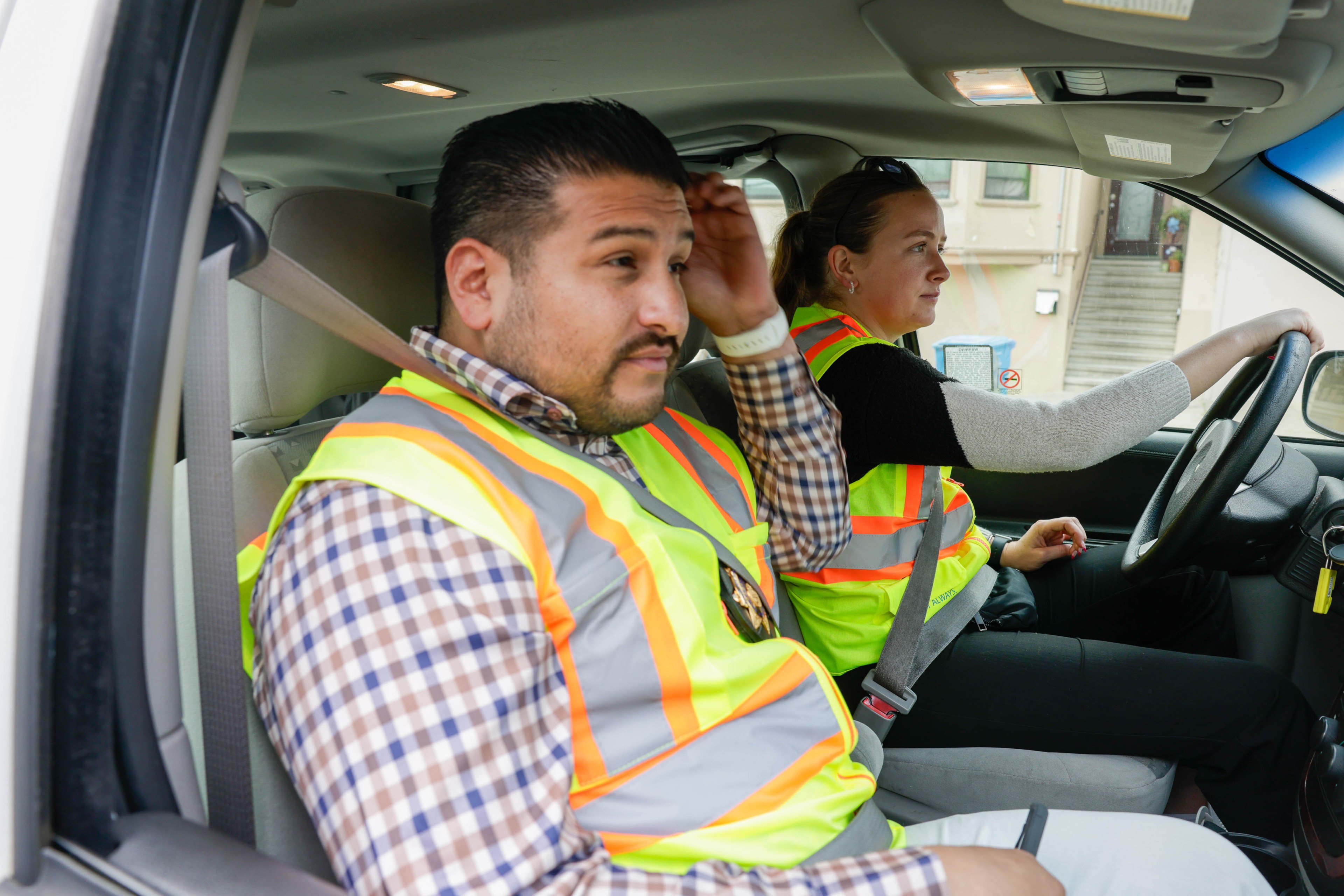 Two people in high-visibility vests are seated in a vehicle; a man in the passenger seat and a woman driving. 