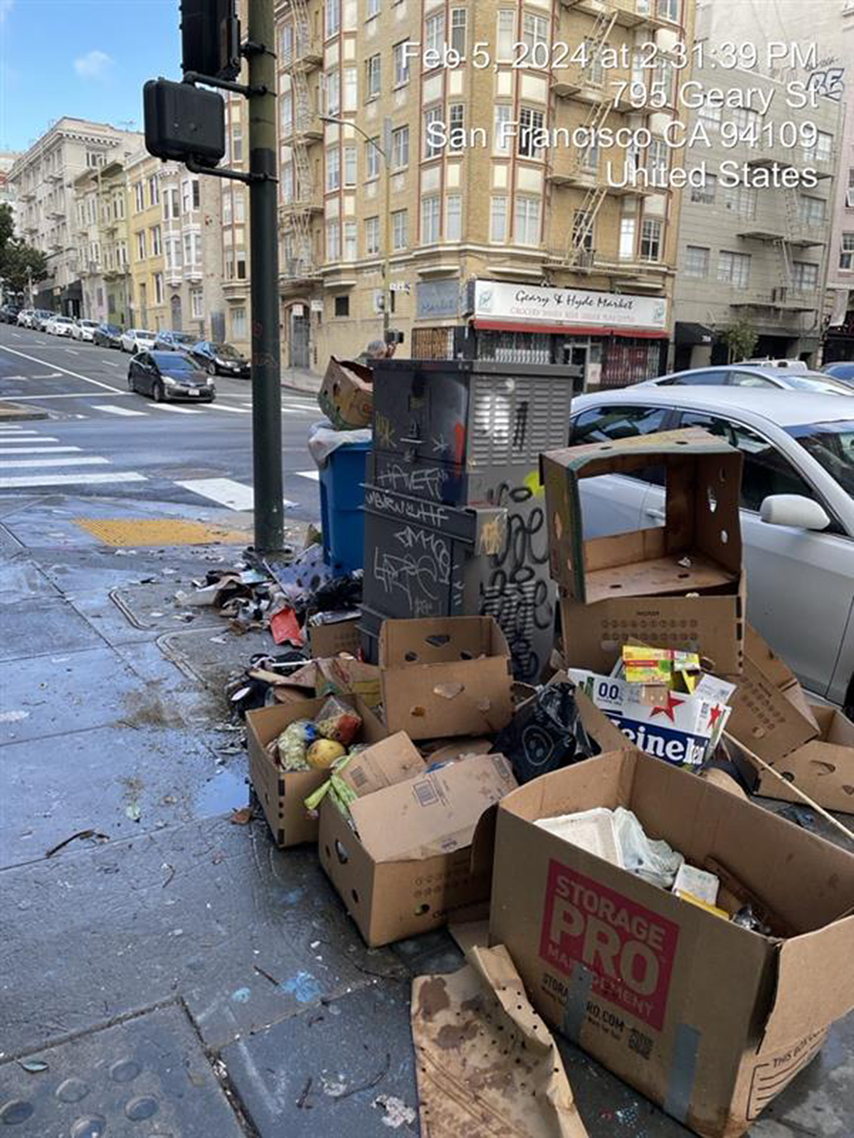 A cluttered city corner with graffiti-covered trash bins surrounded by scattered waste and boxes.