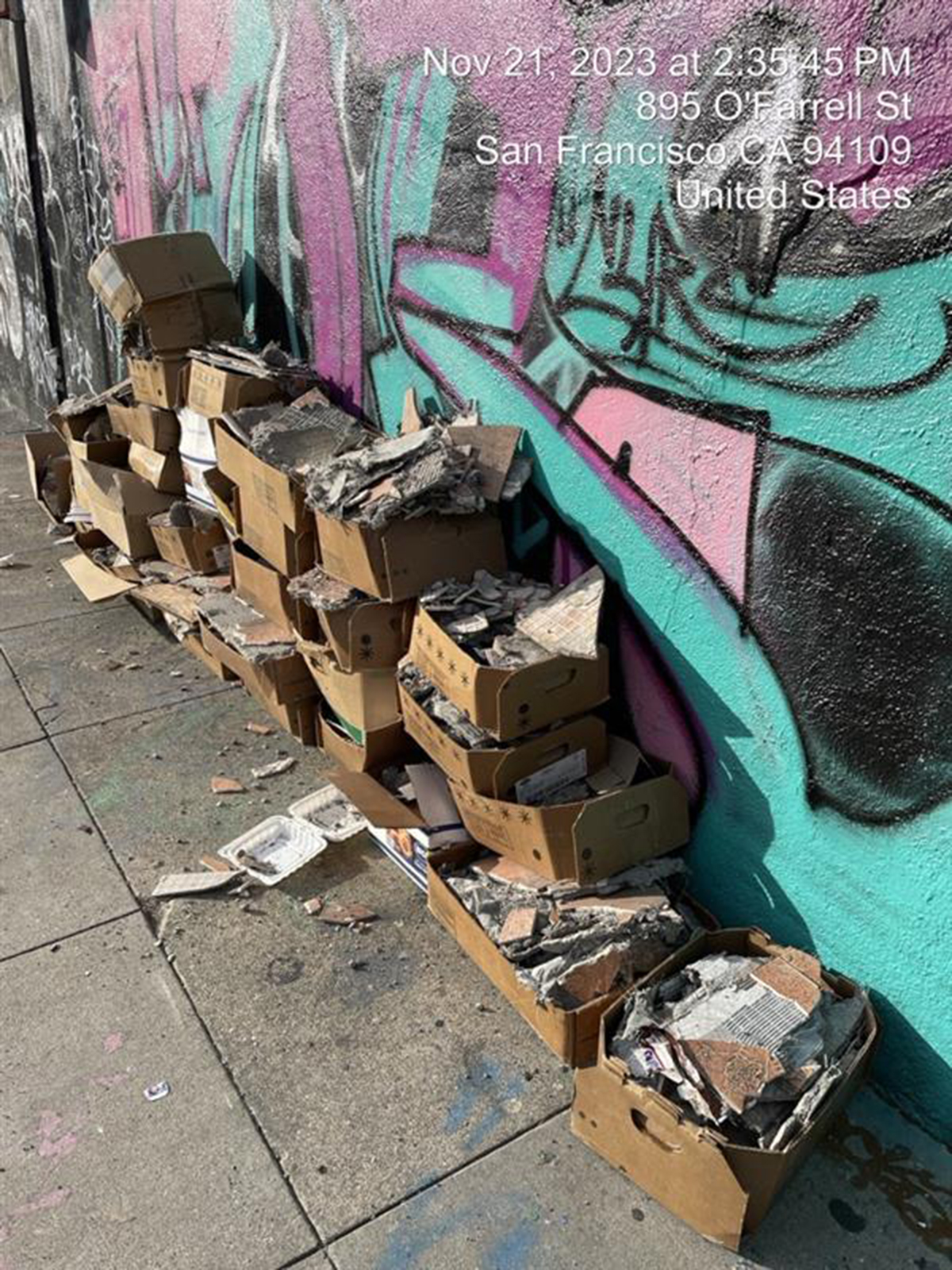 A pile of cardboard boxes filled with debris against a graffiti-covered wall on a sidewalk, timestamped Nov 21, 2023, in San Francisco.