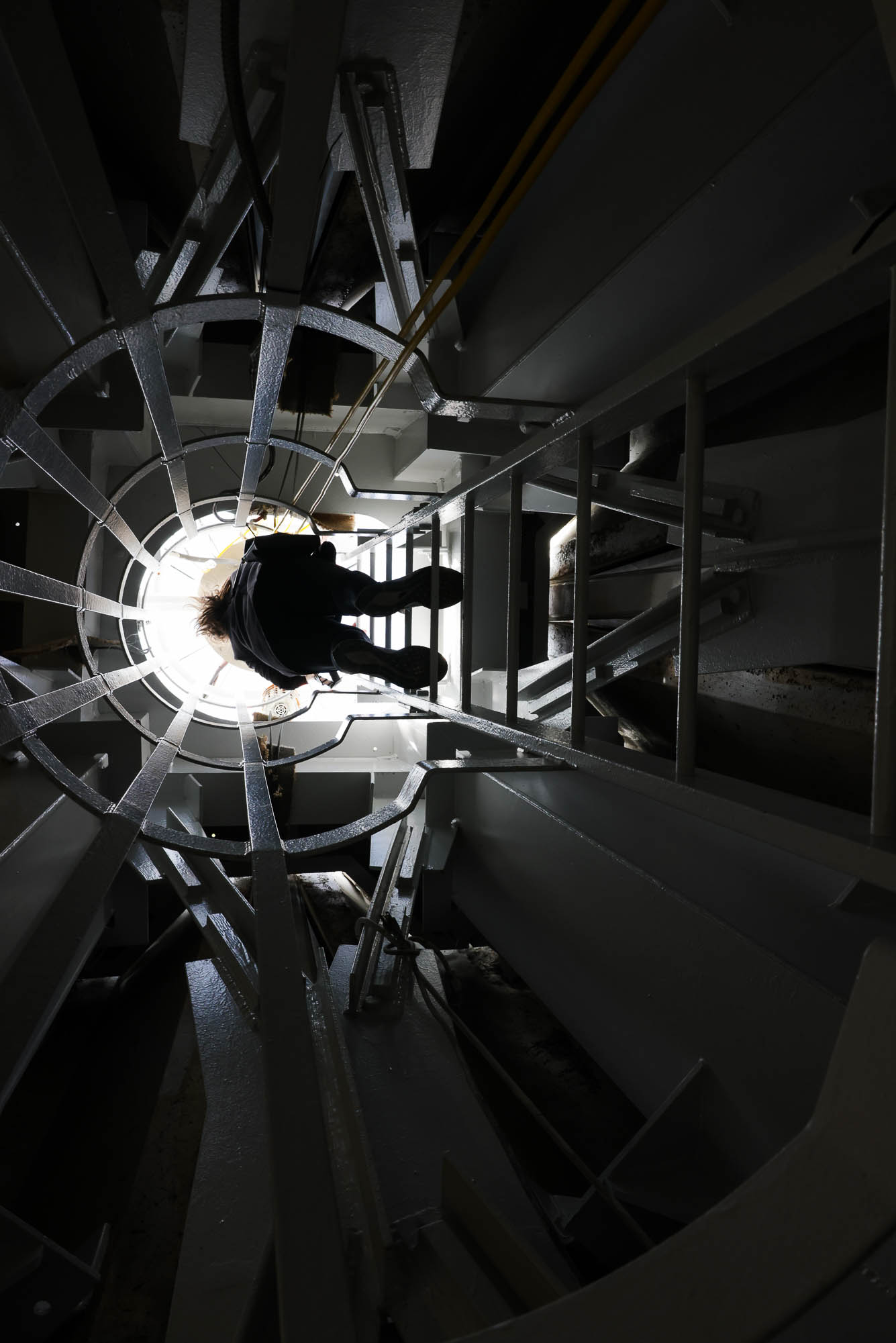 A person ascends a dark, industrial spiral staircase towards a bright light at the top.