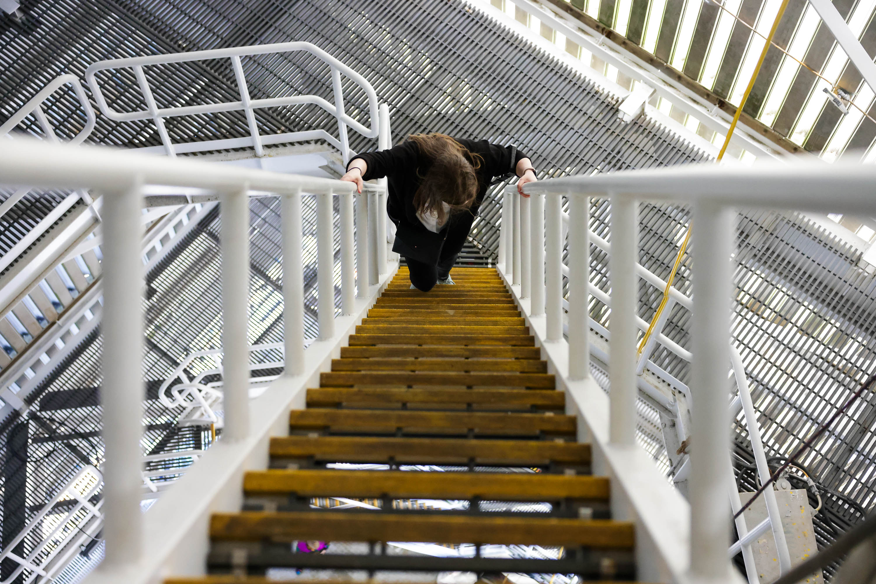 A person is leaning over a white railing at the top of an industrial staircase, looking downwards.