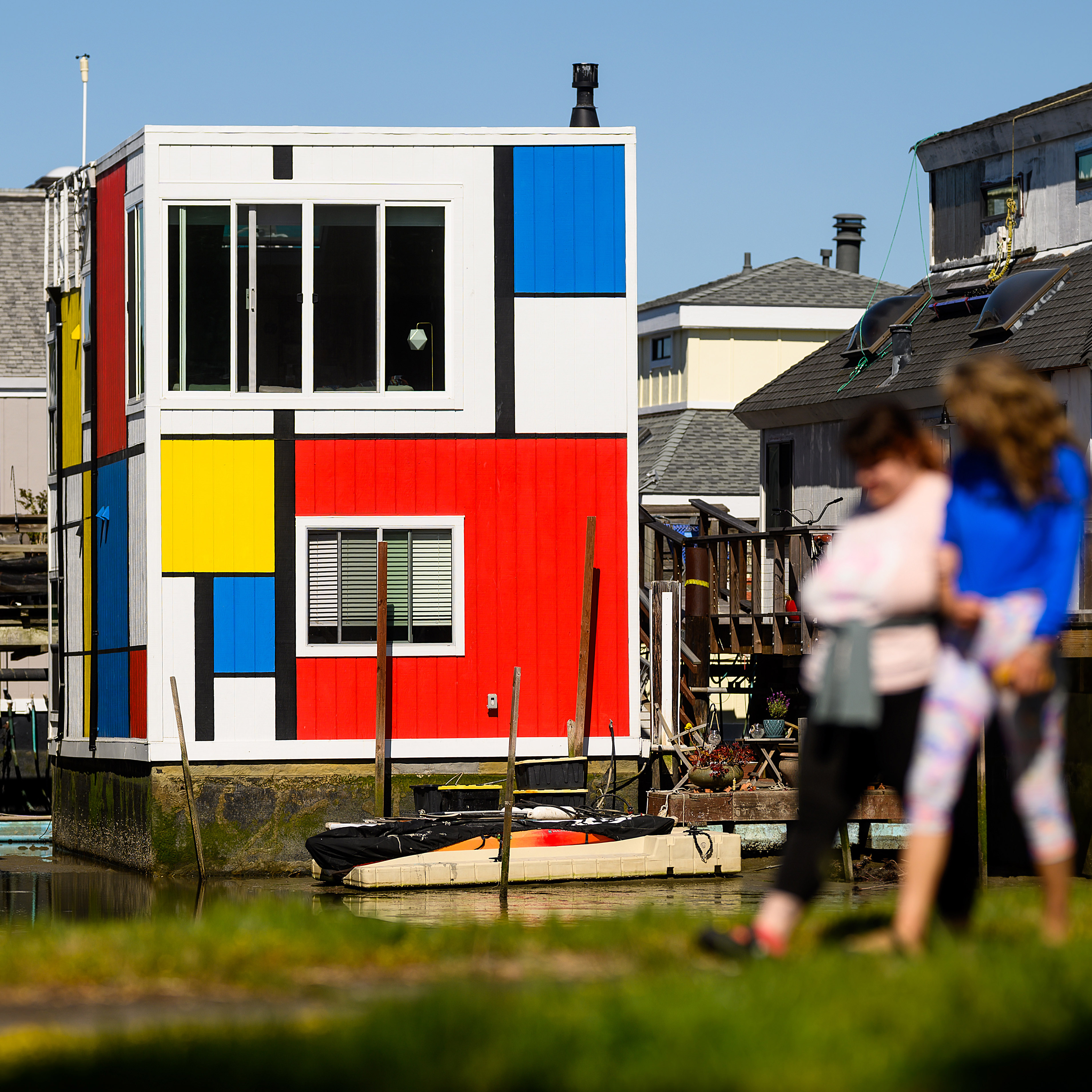 Colorful Mondrian-style house by water, with blurred people walking in front.