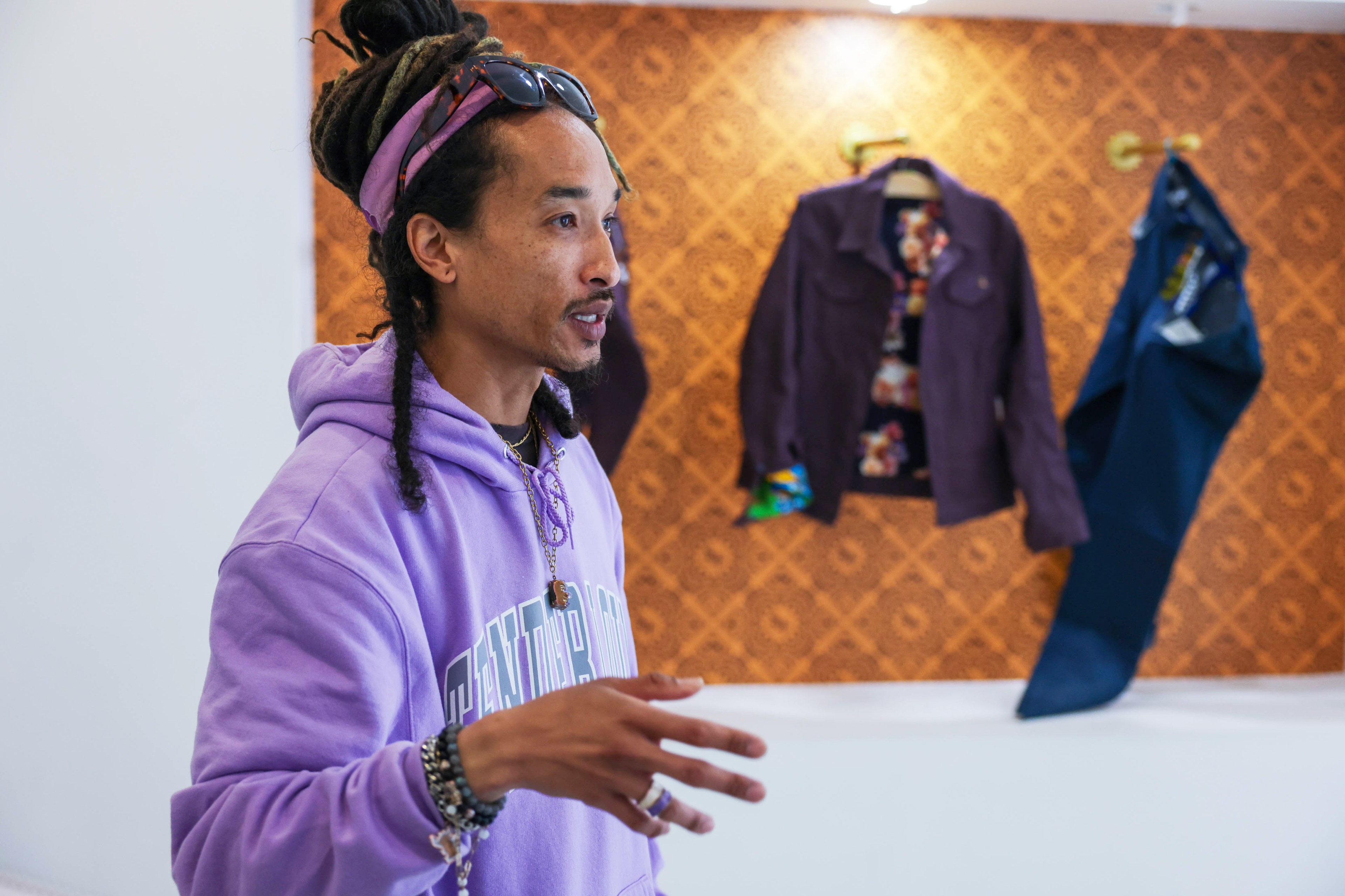 A man in a purple hoodie stands talking, with colorful jackets displayed on a patterned wall behind him.