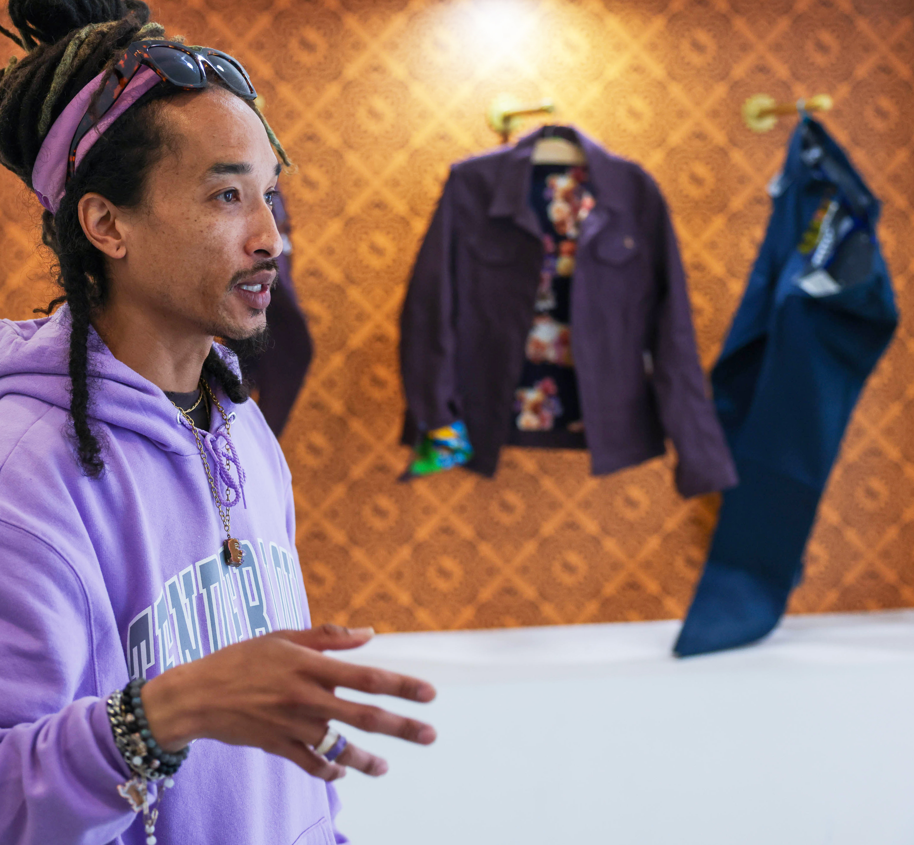 A man with dreadlocks stands in front of a wall displaying colorful jackets.