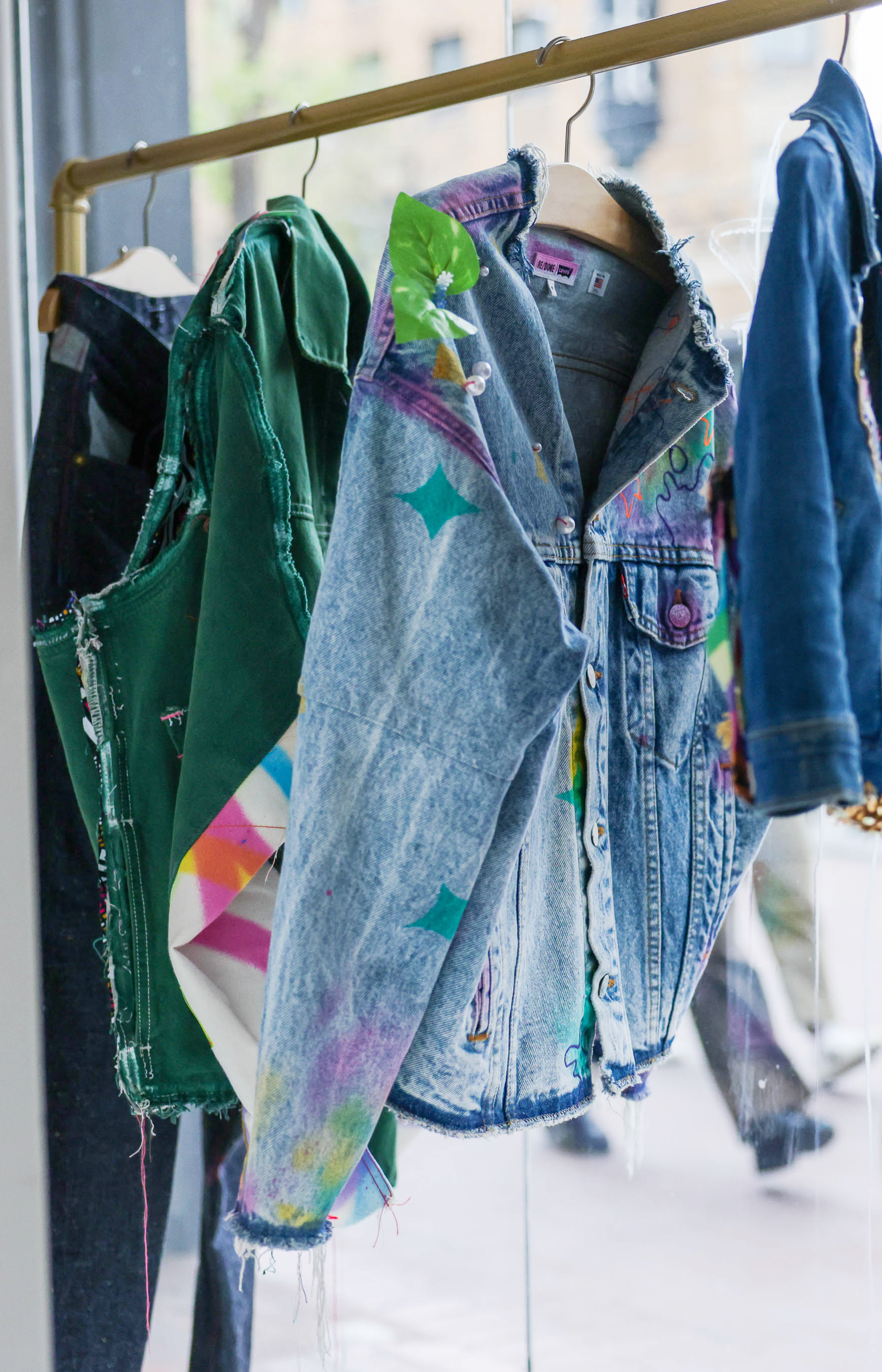 Colorful, distressed denim jackets hang on a rack, with a clear view through a window behind.