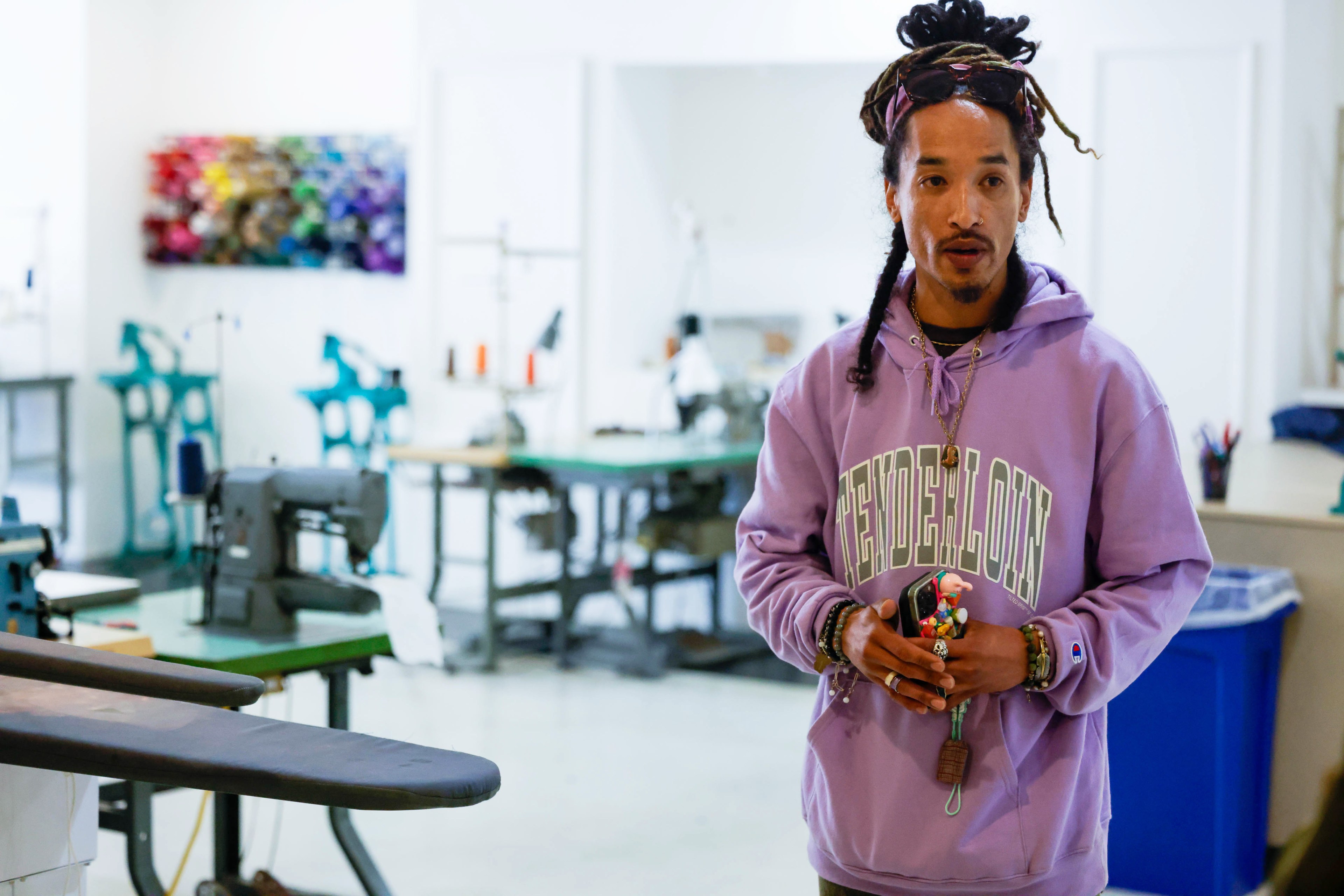 A man stands in a workshop with sewing machines, wearing a purple hoodie and adorned with jewelry.