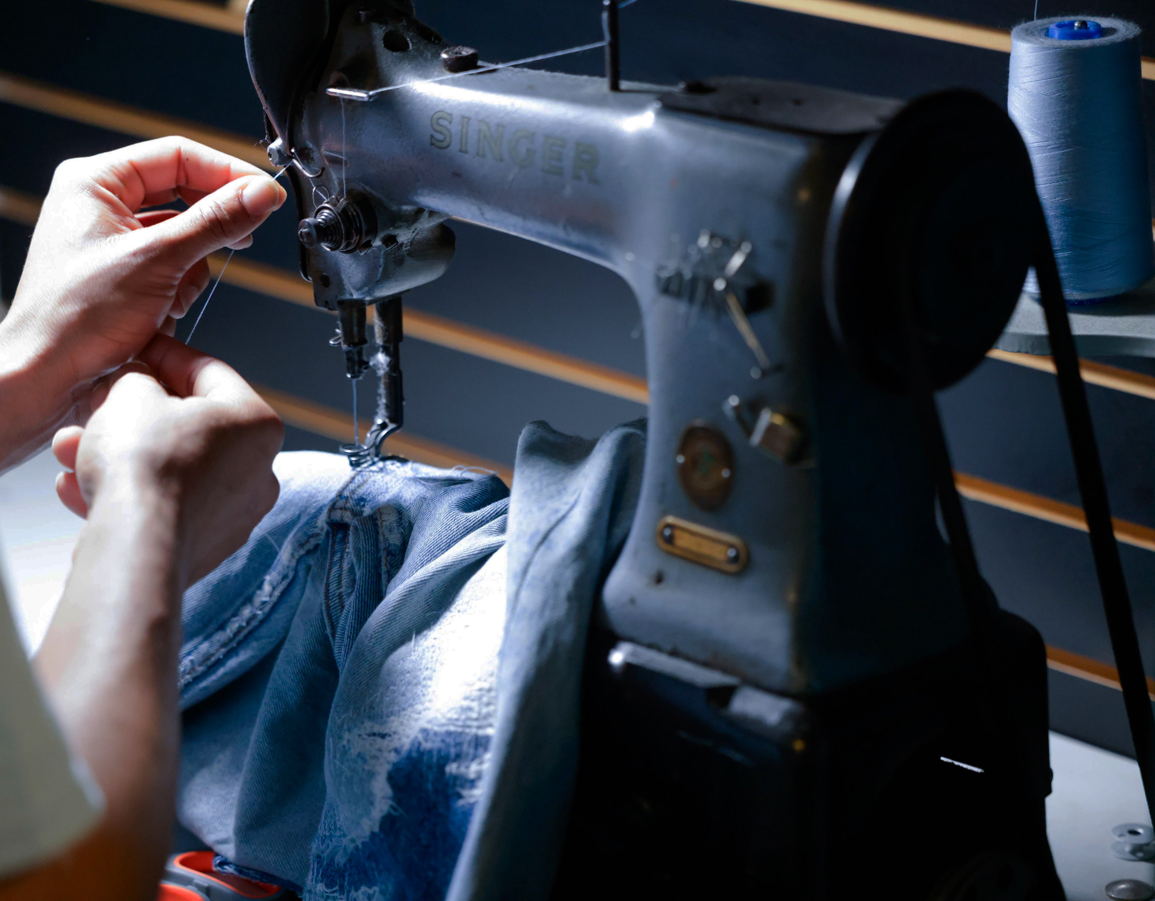 Hands threading a needle on a vintage Singer sewing machine with denim and blue thread spool.