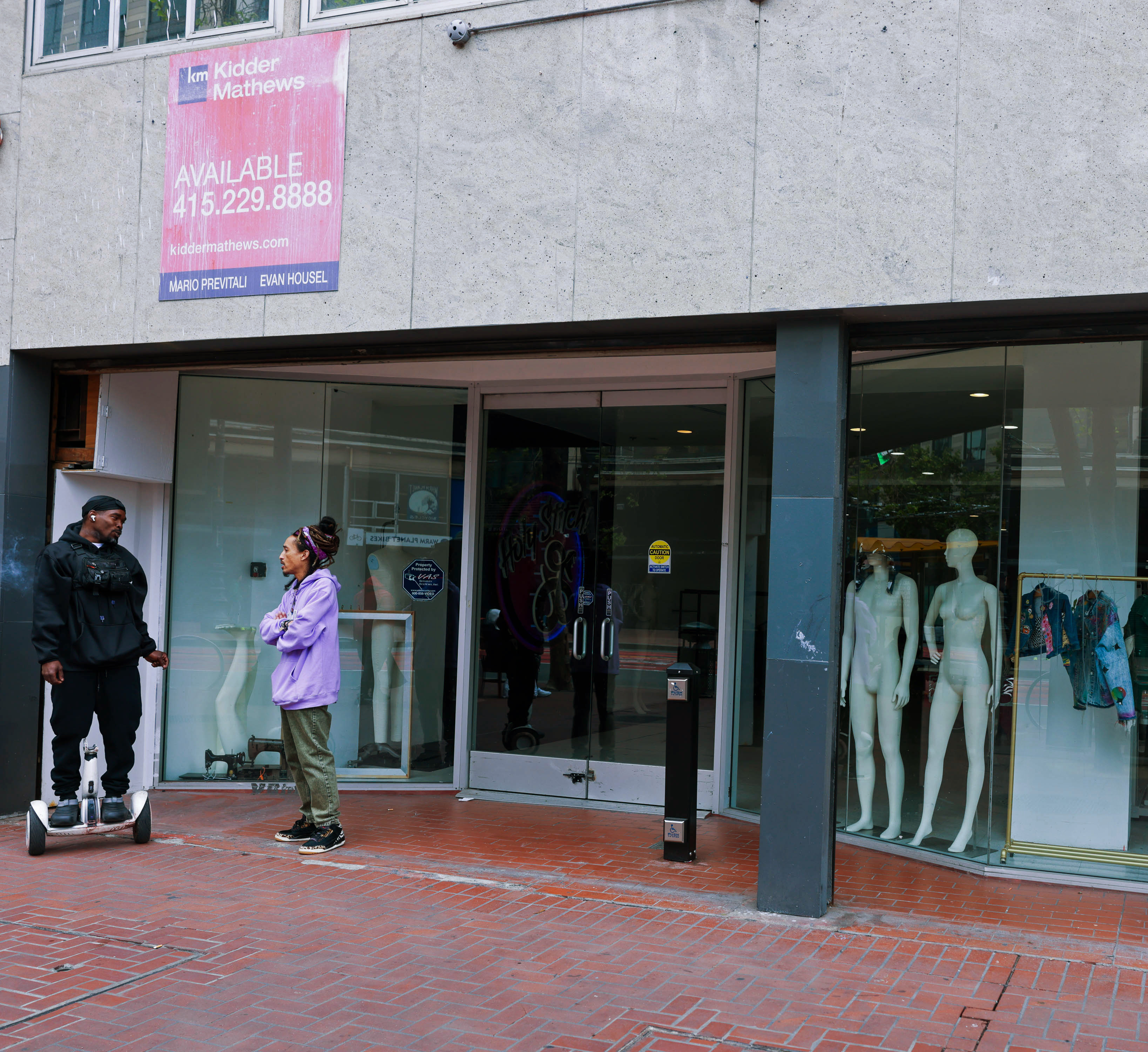 Two people converse outside a store with mannequins and a 'For Lease' sign above.