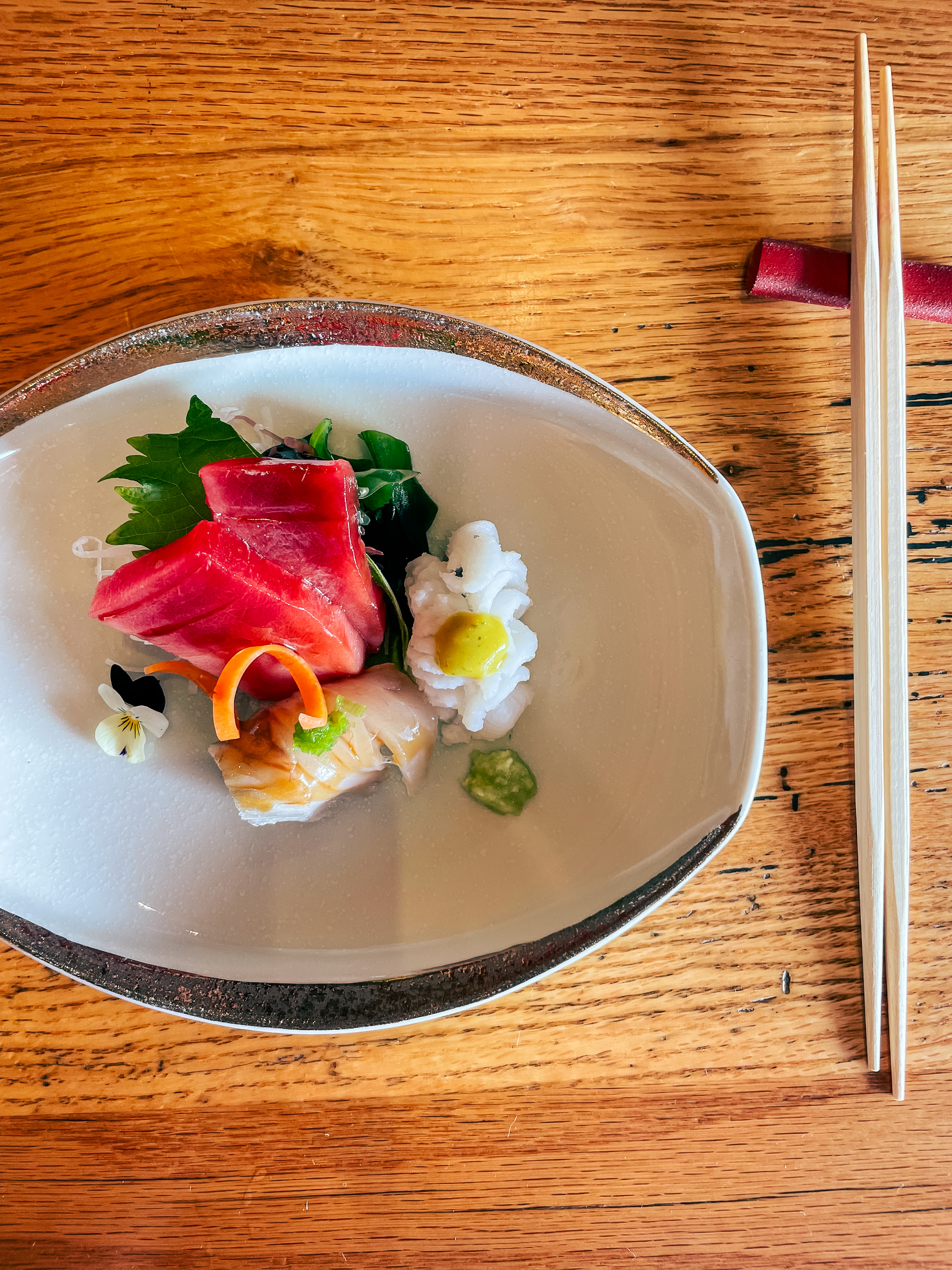 The image shows a beautifully plated dish of sashimi, including tuna, white fish, and squid, garnished with a yellow dollop, a green paste, a small flower, and decorative vegetables. It is presented on an oval-shaped plate next to a pair of chopsticks on a wooden table.