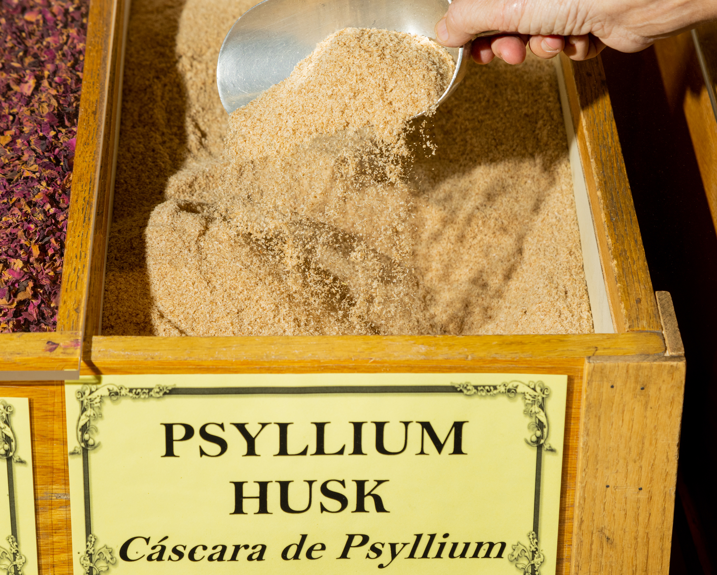 A hand is scooping psyllium husk from a wooden bin labeled &quot;PSYLLIUM HUSK Cáscara de Psyllium.&quot;