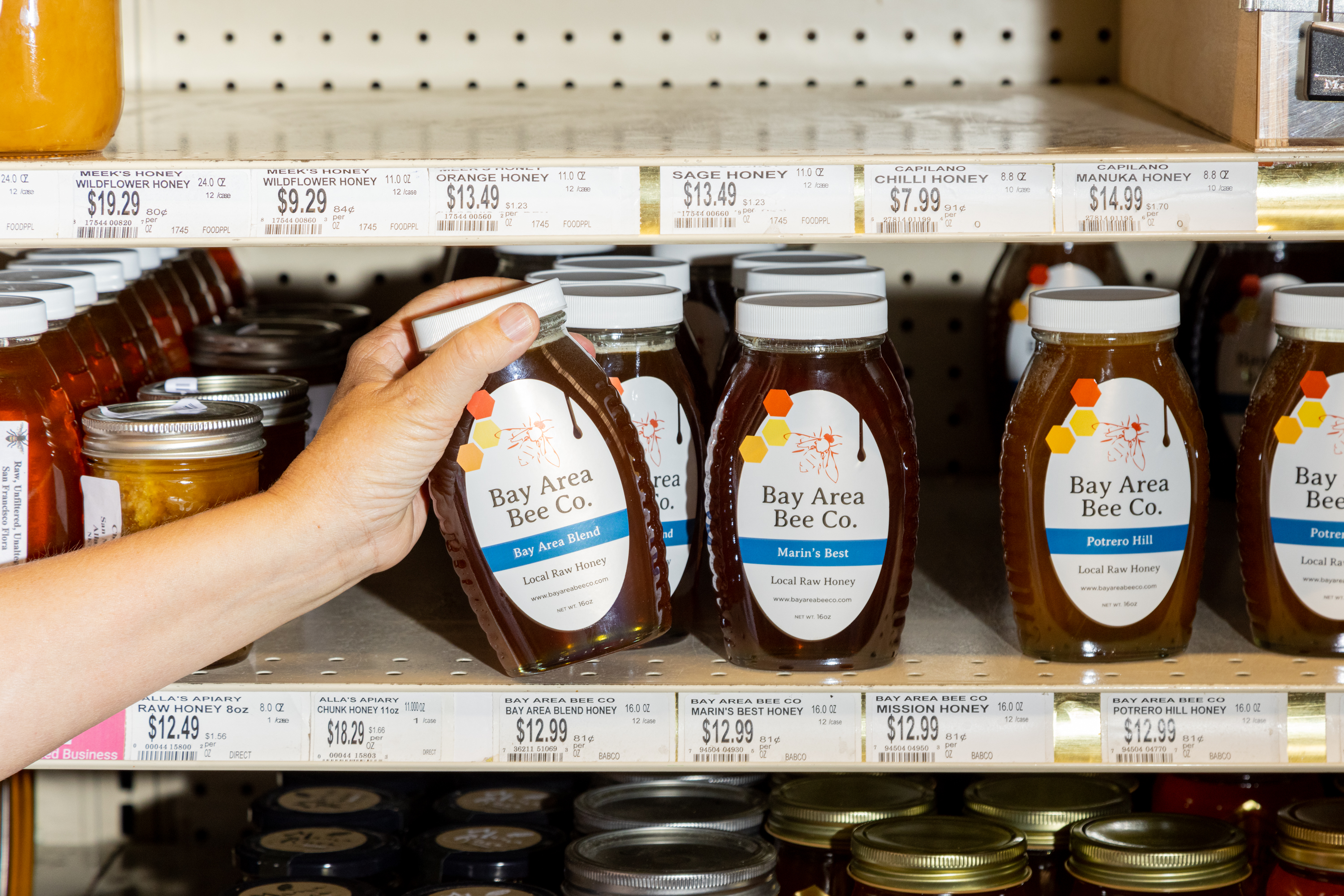 A person's hand is selecting a jar labeled &quot;Bay Area Bee Co.&quot; from a shelf of various honeys at a store.