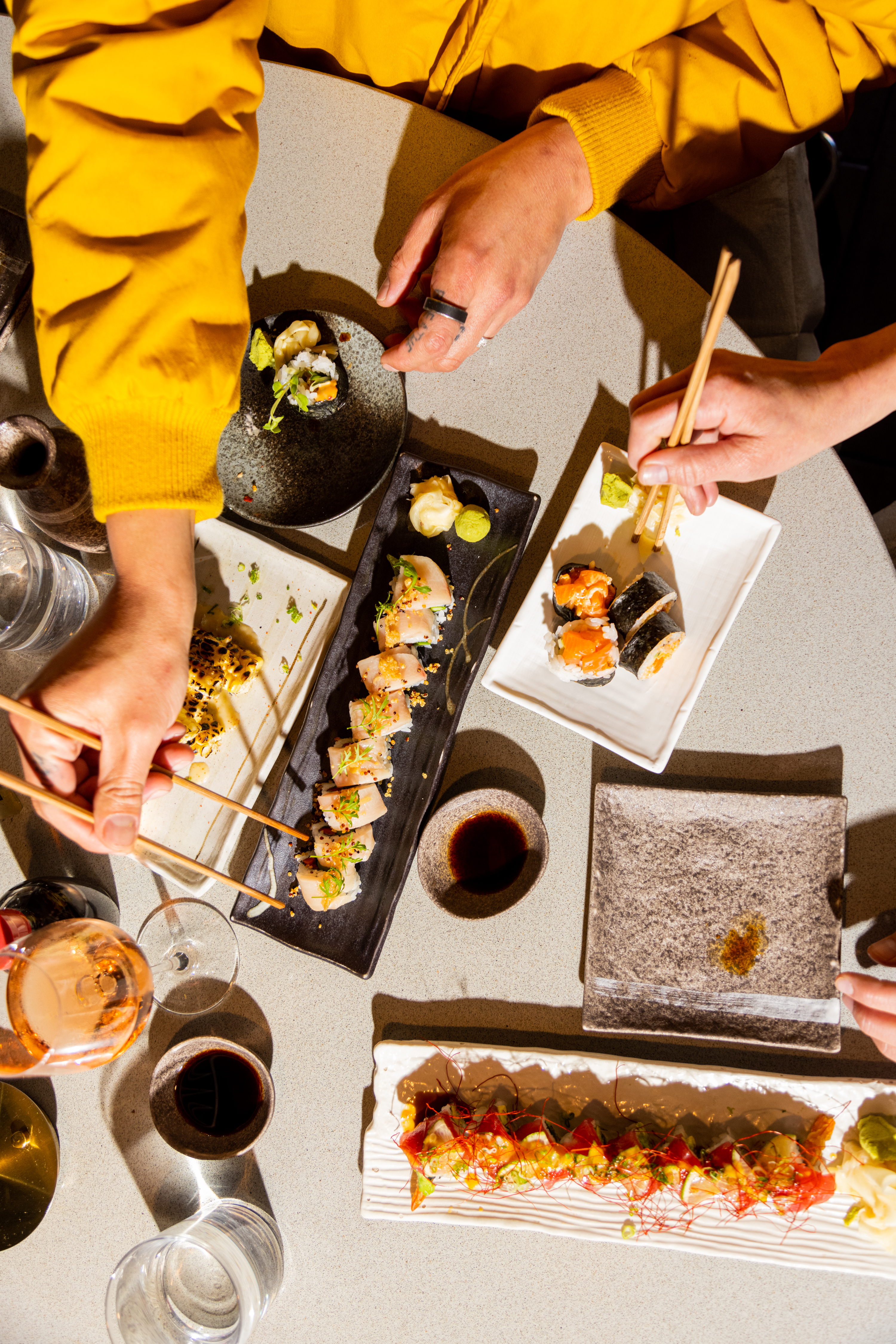 Two people are using chopsticks to eat various sushi dishes arranged on a table, including sushi rolls and nigiri, with dipping sauces and beverages.
