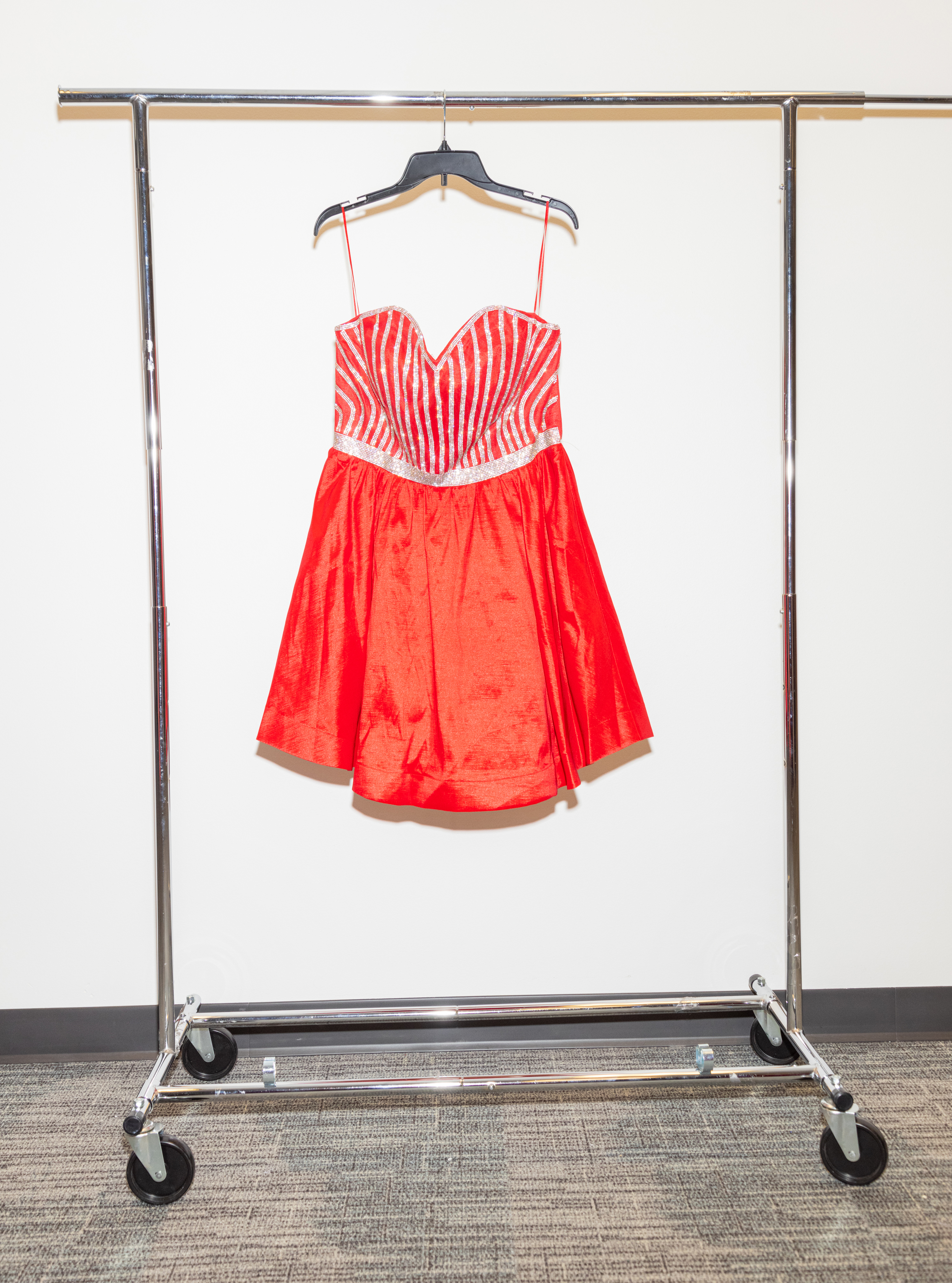 A red, strapless dress with white stripes and rhinestone accents, hanging on a rack against a white wall.