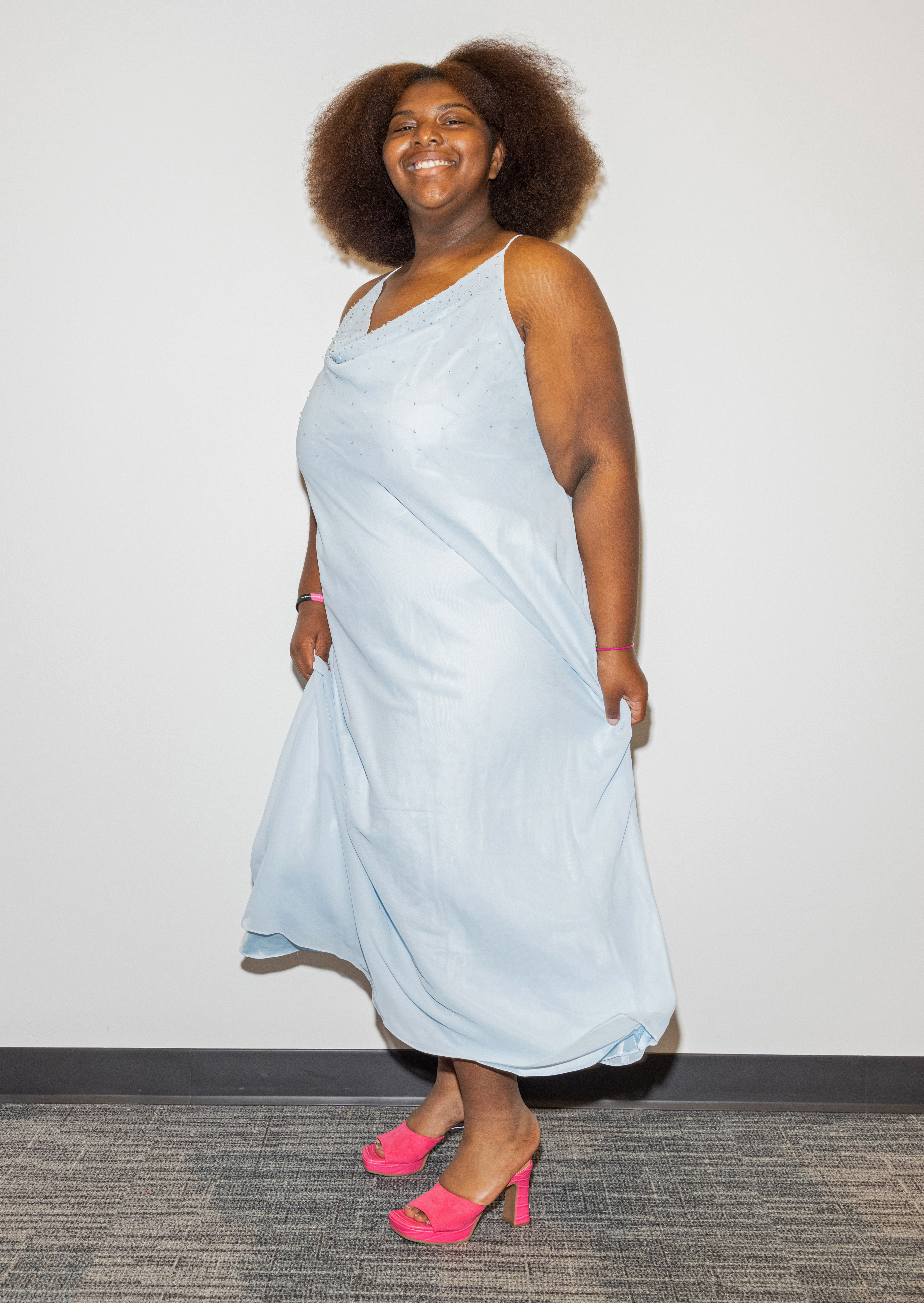 A smiling woman in a light blue dress and pink high heels, with her voluminous hair styled in an afro.