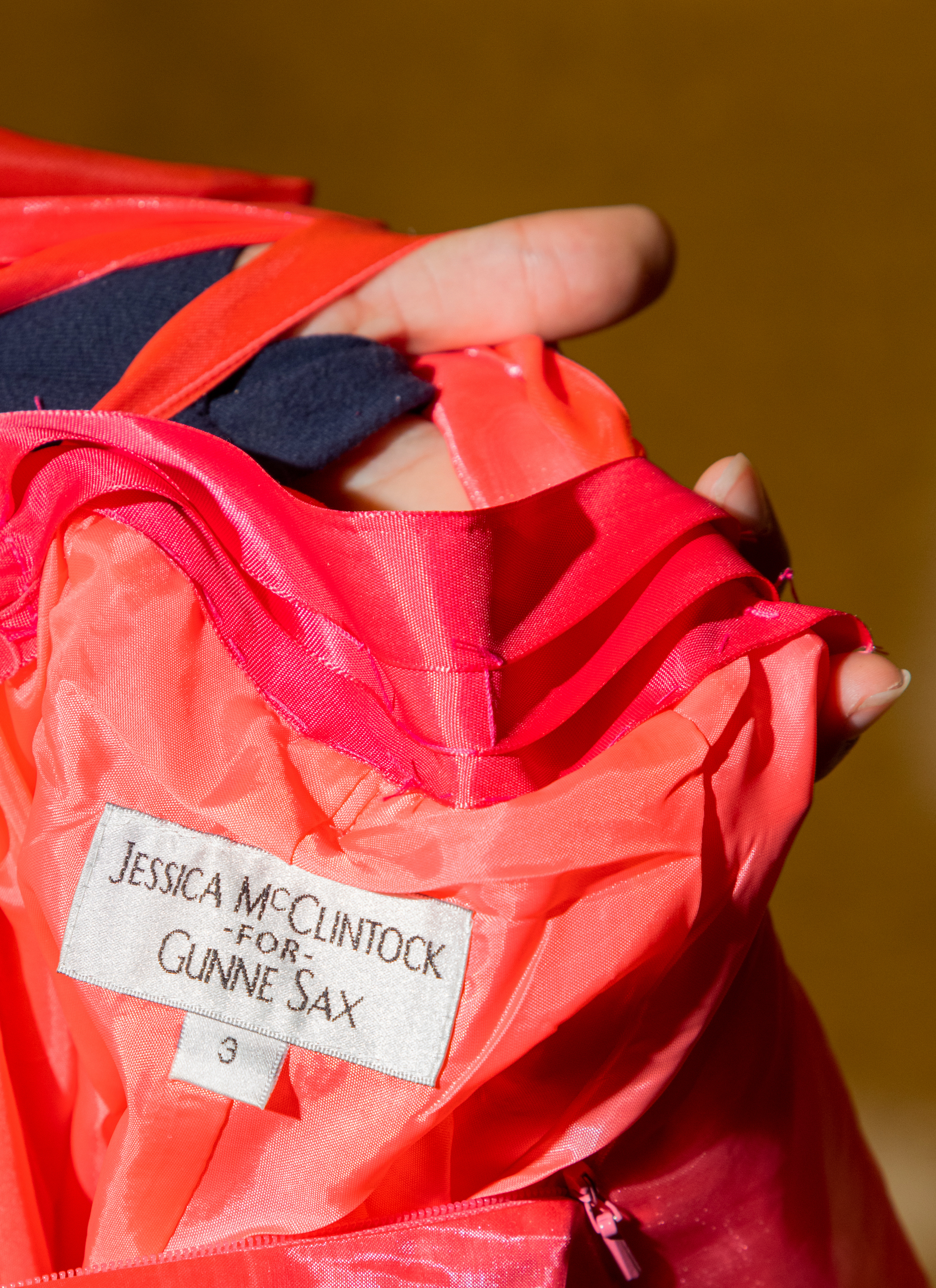 A label on a red fabric reads &quot;Jessica McClintock for Gunne Sax&quot; with a hand partially covering it.