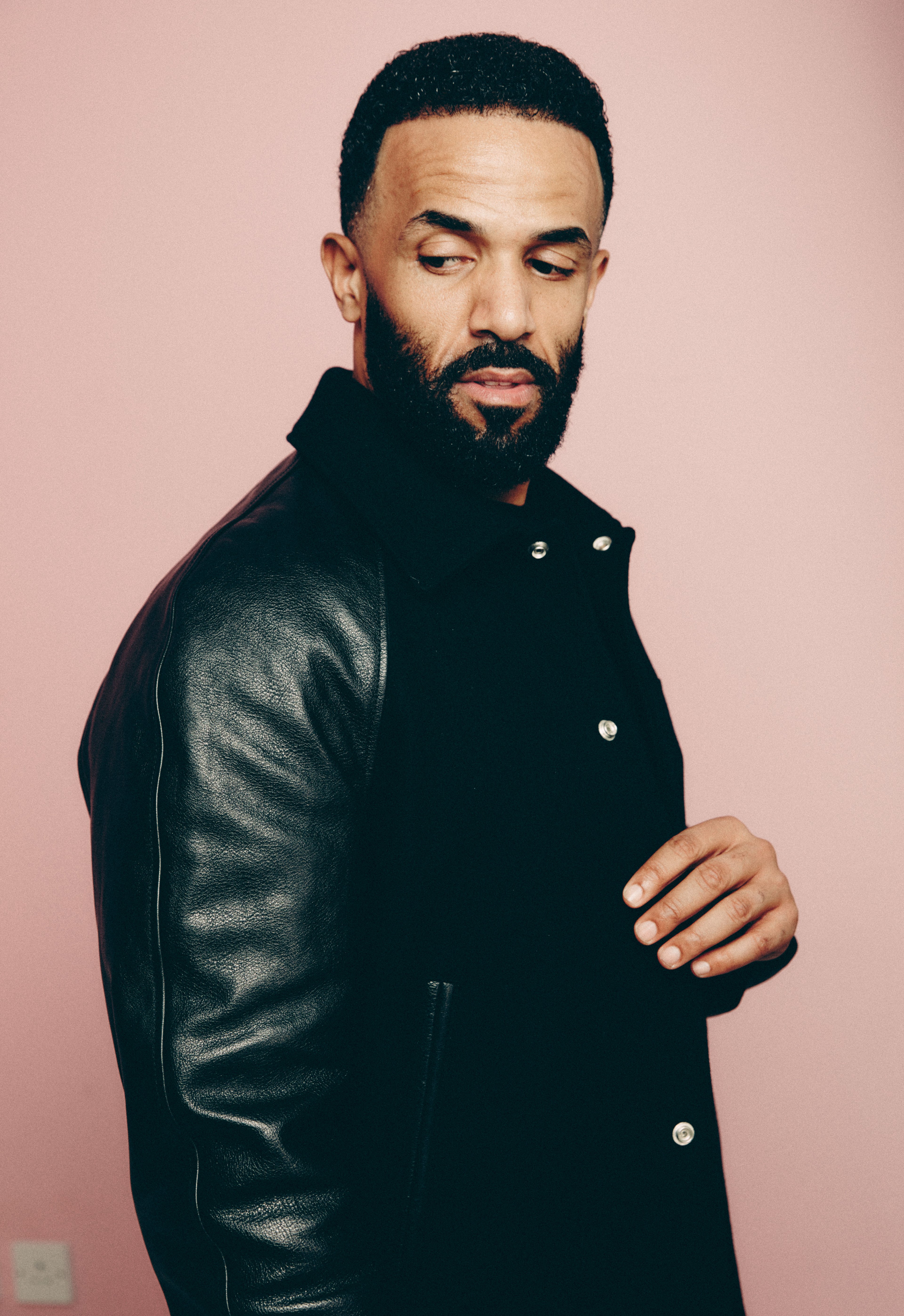 A man with a neat beard, wearing a black jacket, stands before a pink background. He is holding the jacket and gazing at the camera.