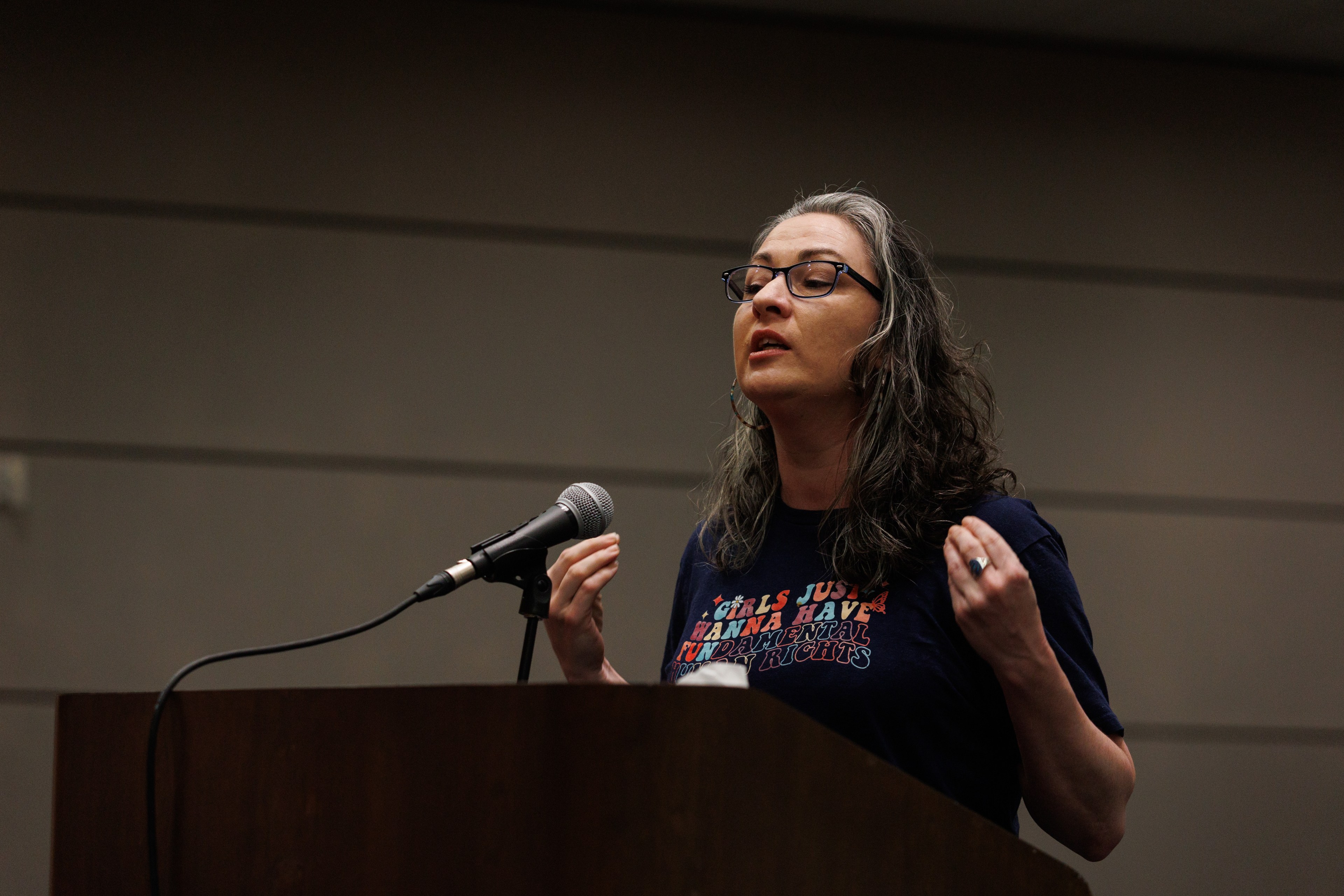 A woman with glasses is speaking into a microphone at a podium, gesturing with her hands.