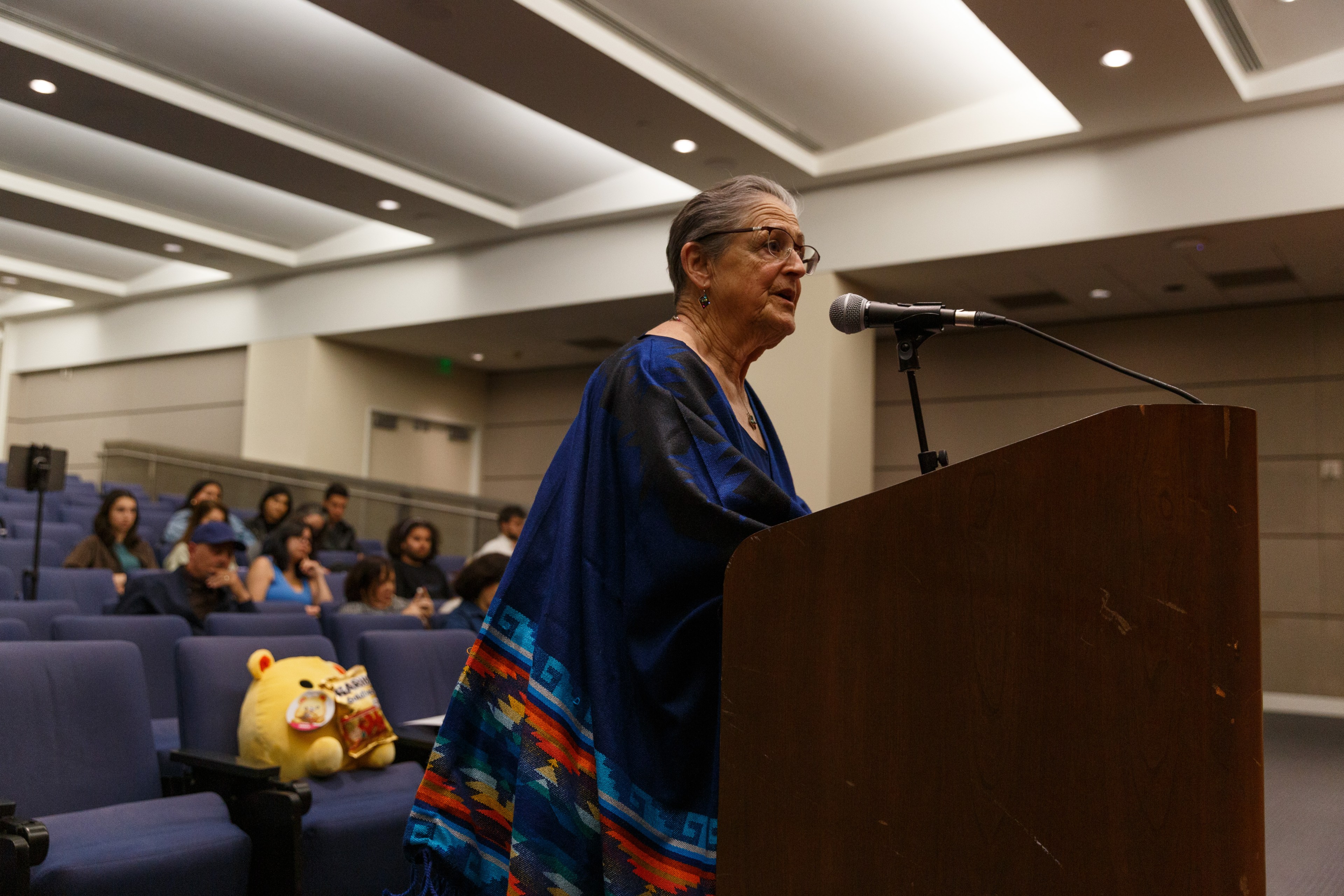 An elderly woman speaks at a podium in a lecture hall with an audience behind her.