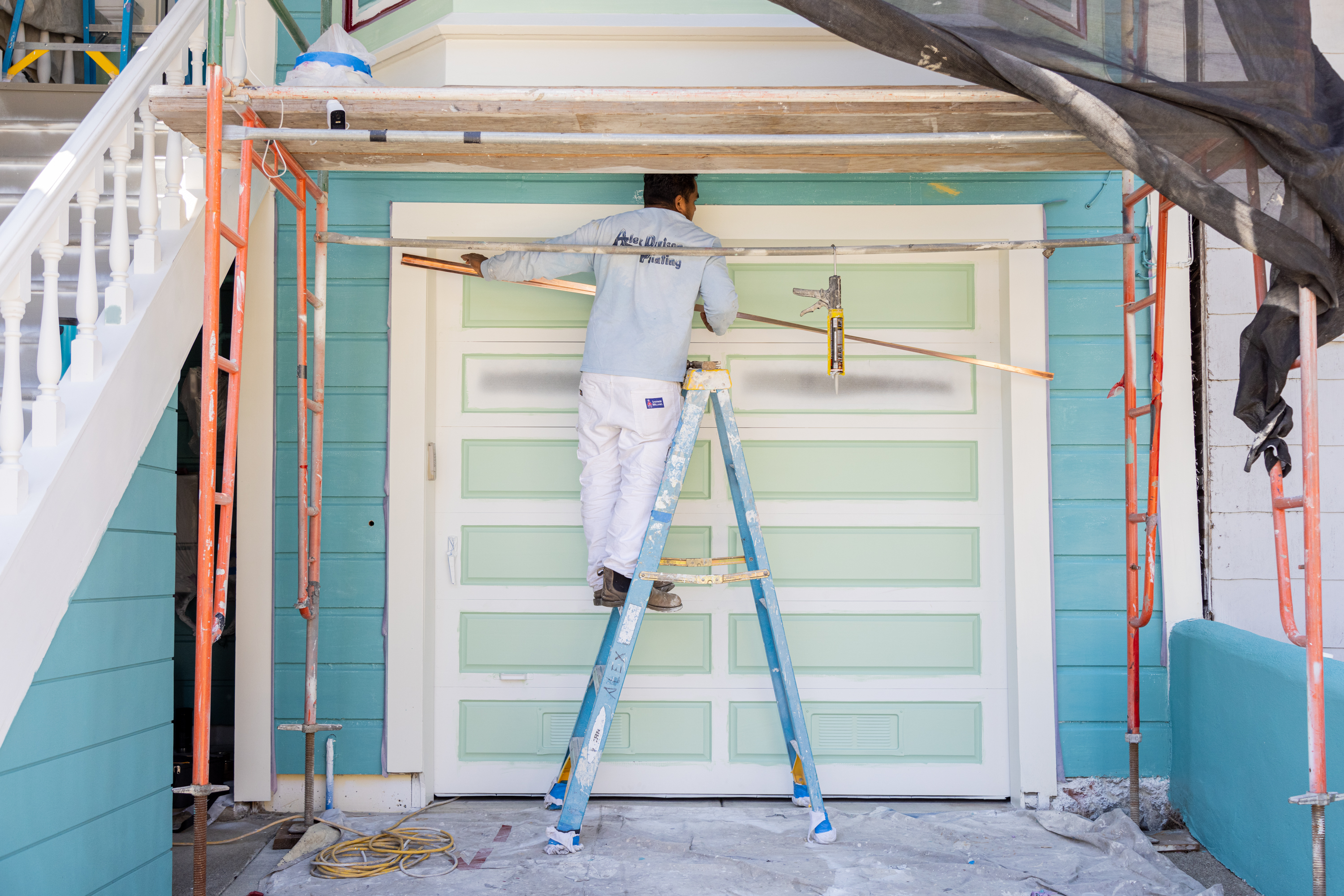 A person on a ladder is installing trim above a green garage door, surrounded by construction tools and materials.