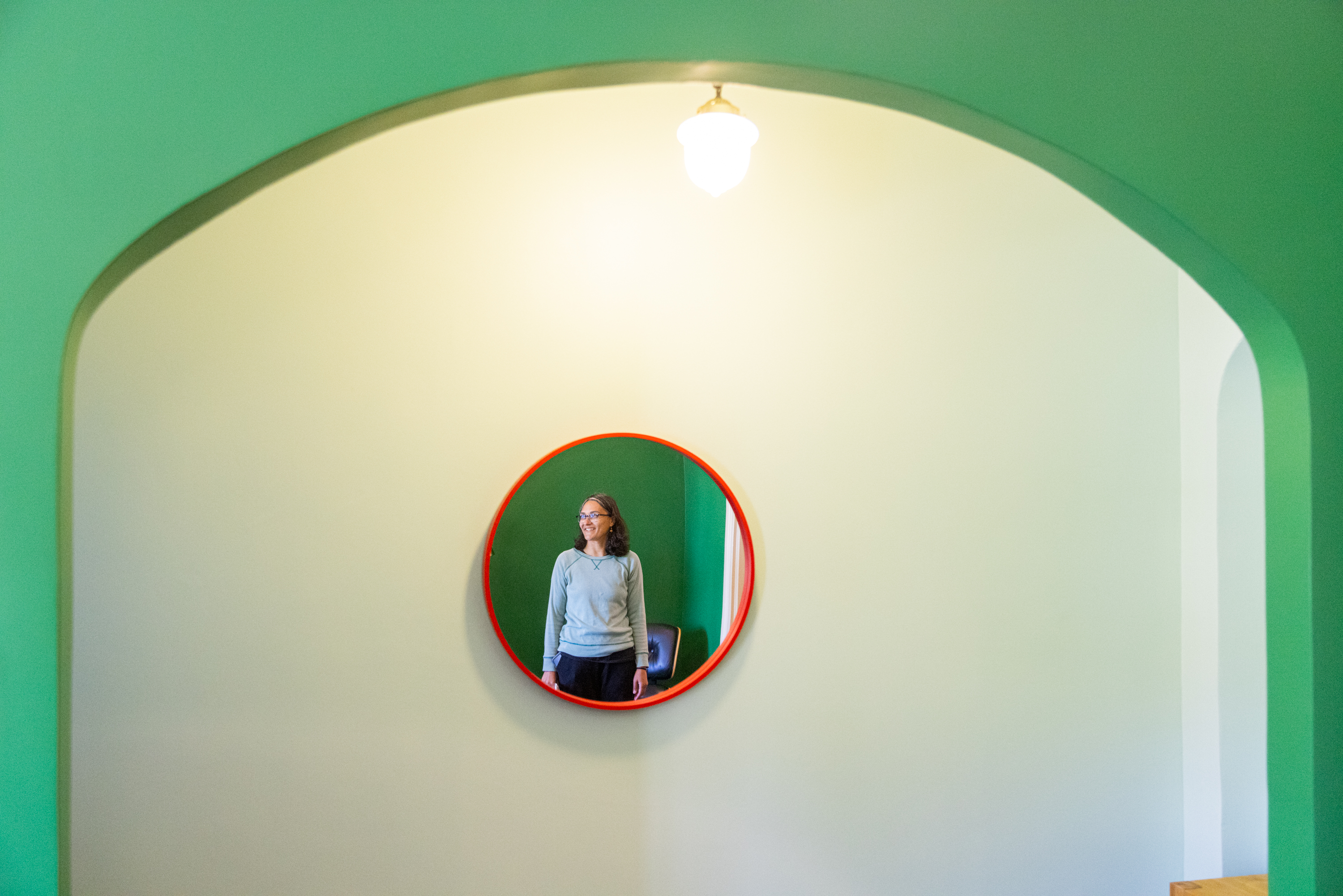 A woman is reflected in a round mirror with a red frame on a green and cream wall, under an arched alcove with a hanging light.