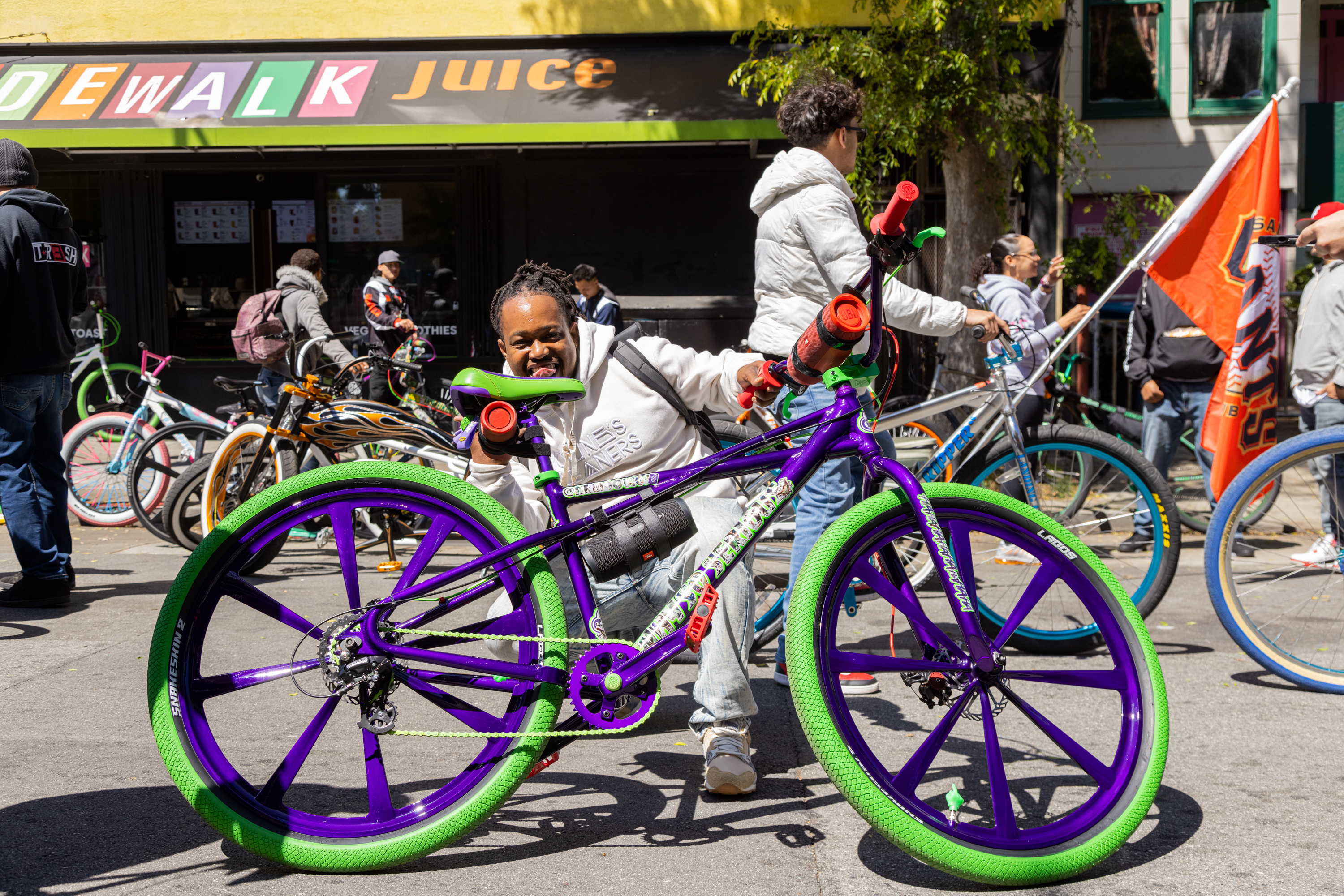 A person smiles behind a custom bike with purple frame and green wheels on a sunny street with onlookers and bikes.