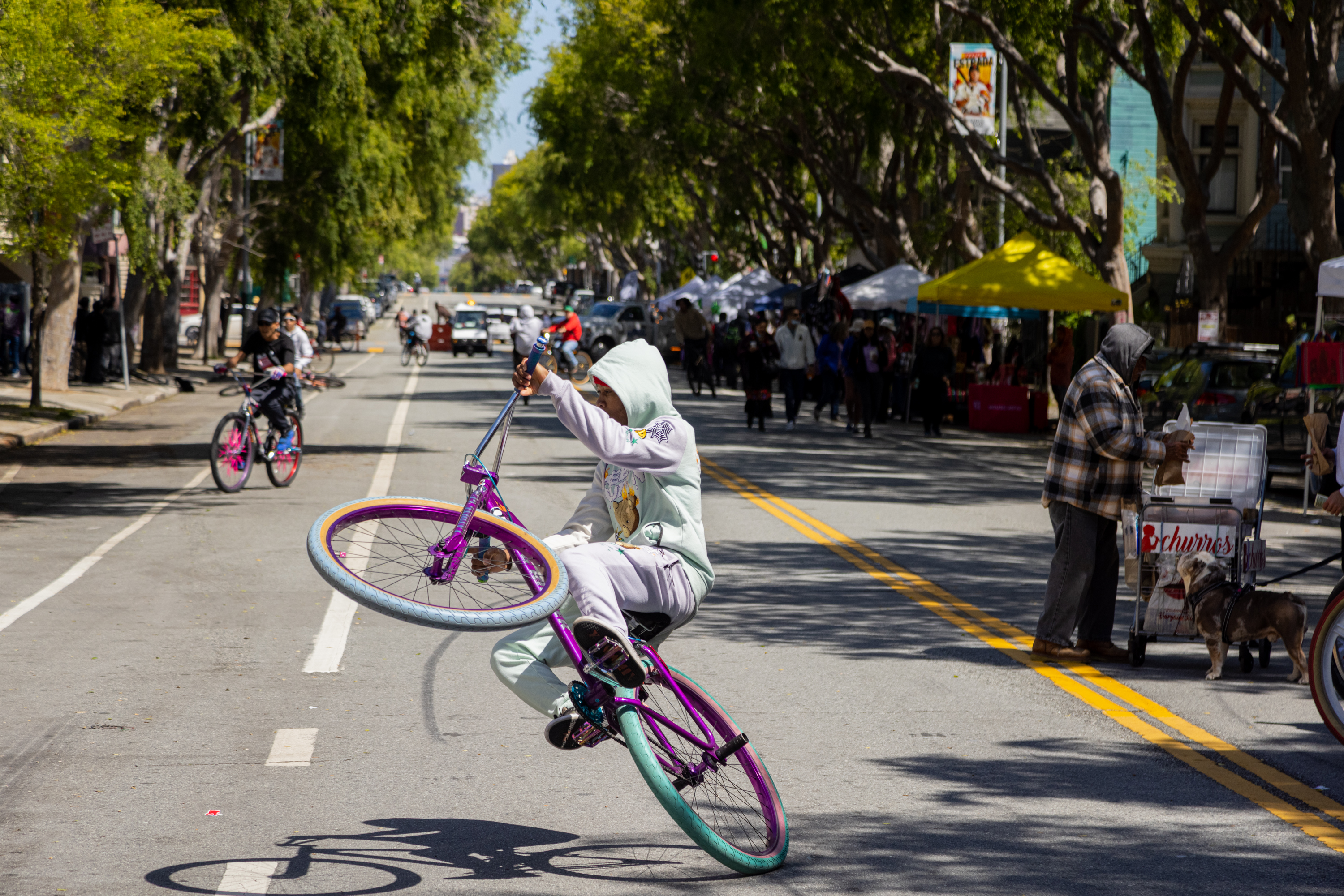 A person is performing a wheelie on a colorful bike down a sunny, tree-lined street with bystanders and stalls.