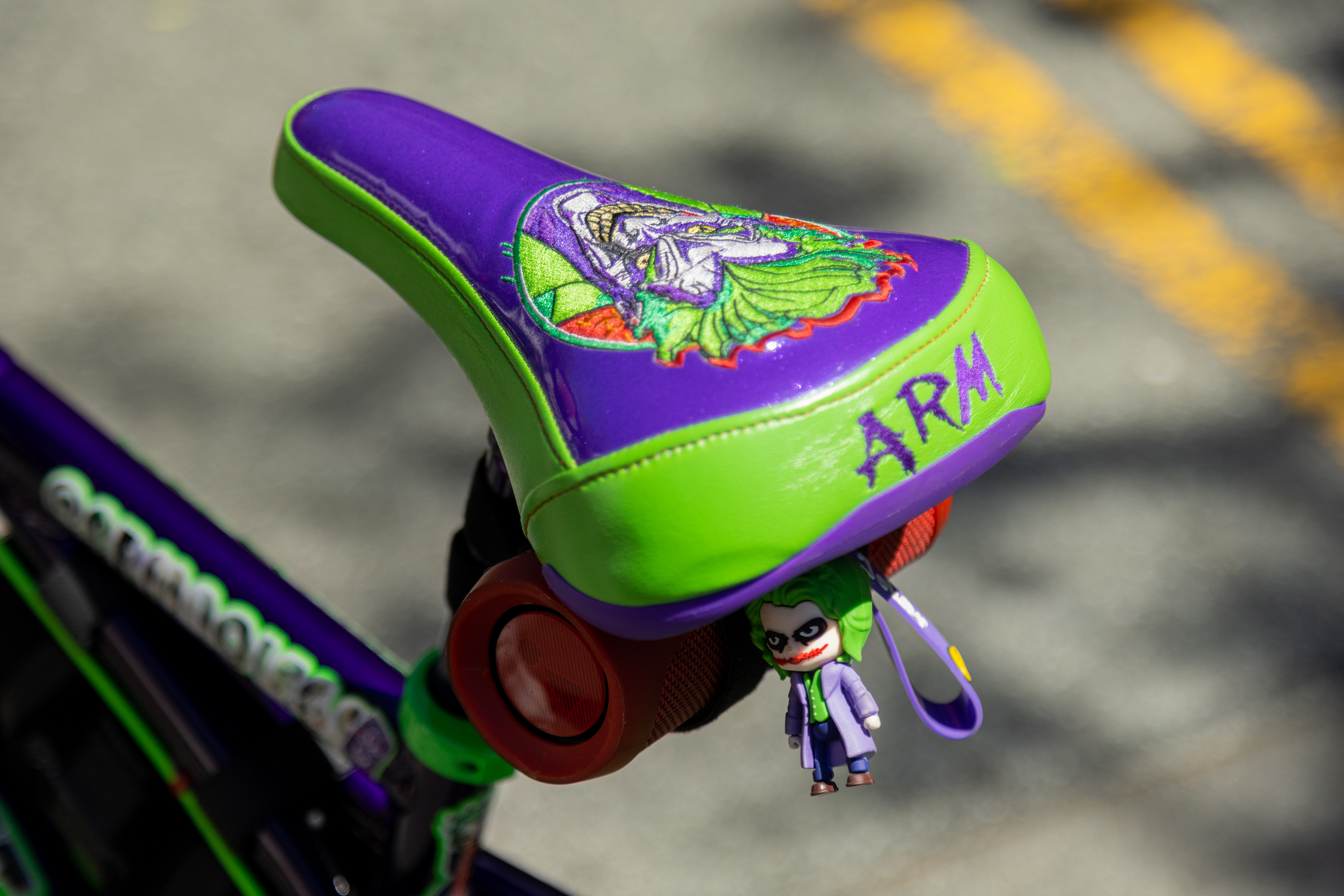 A vibrant purple and green bike saddle with an emblem and the word &quot;ARM&quot; on it, with a small Joker figure hanging below.
