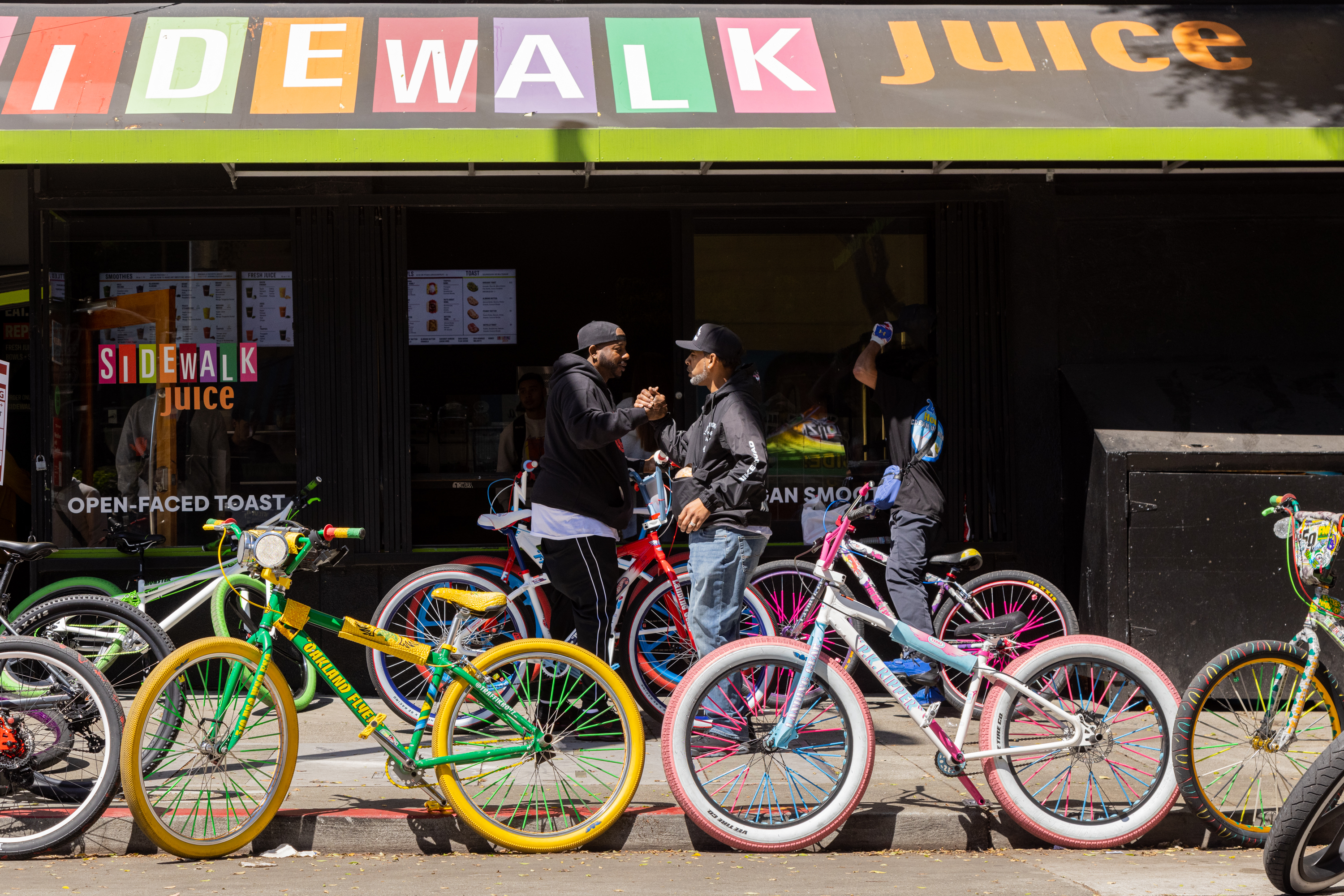 Two people greet each other in front of a colorful juice bar with vibrant custom bikes parked outside.