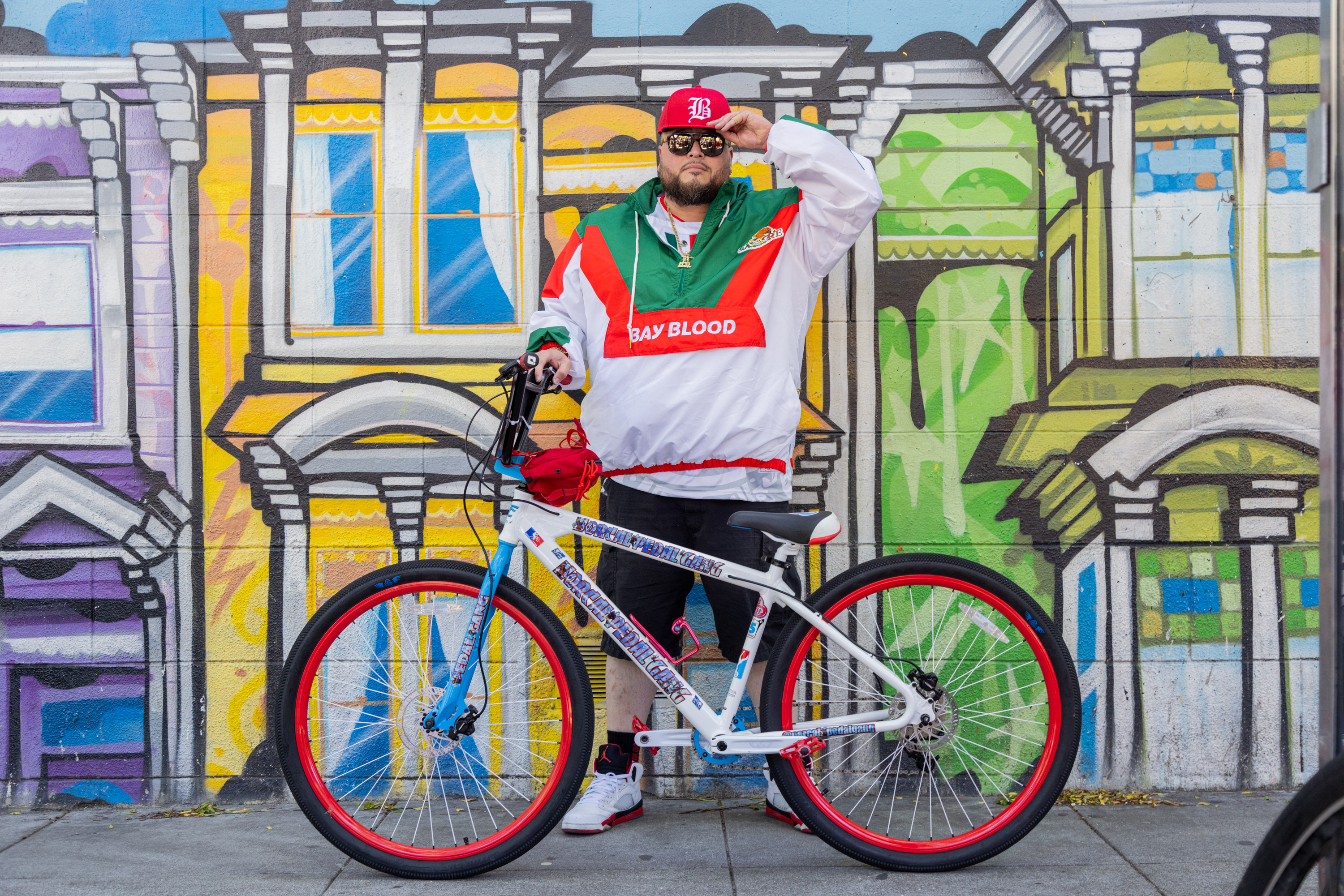 A man stands with a bike before a colorful mural dressed in sporty attire, exuding a cool urban vibe.