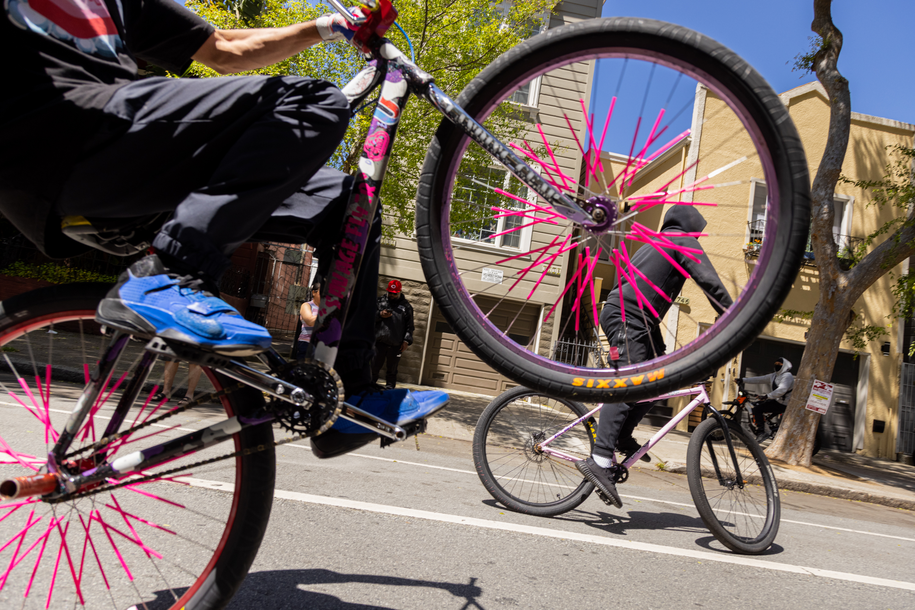 Cyclists perform stunts on bikes with vibrant pink spokes on a sunny street.