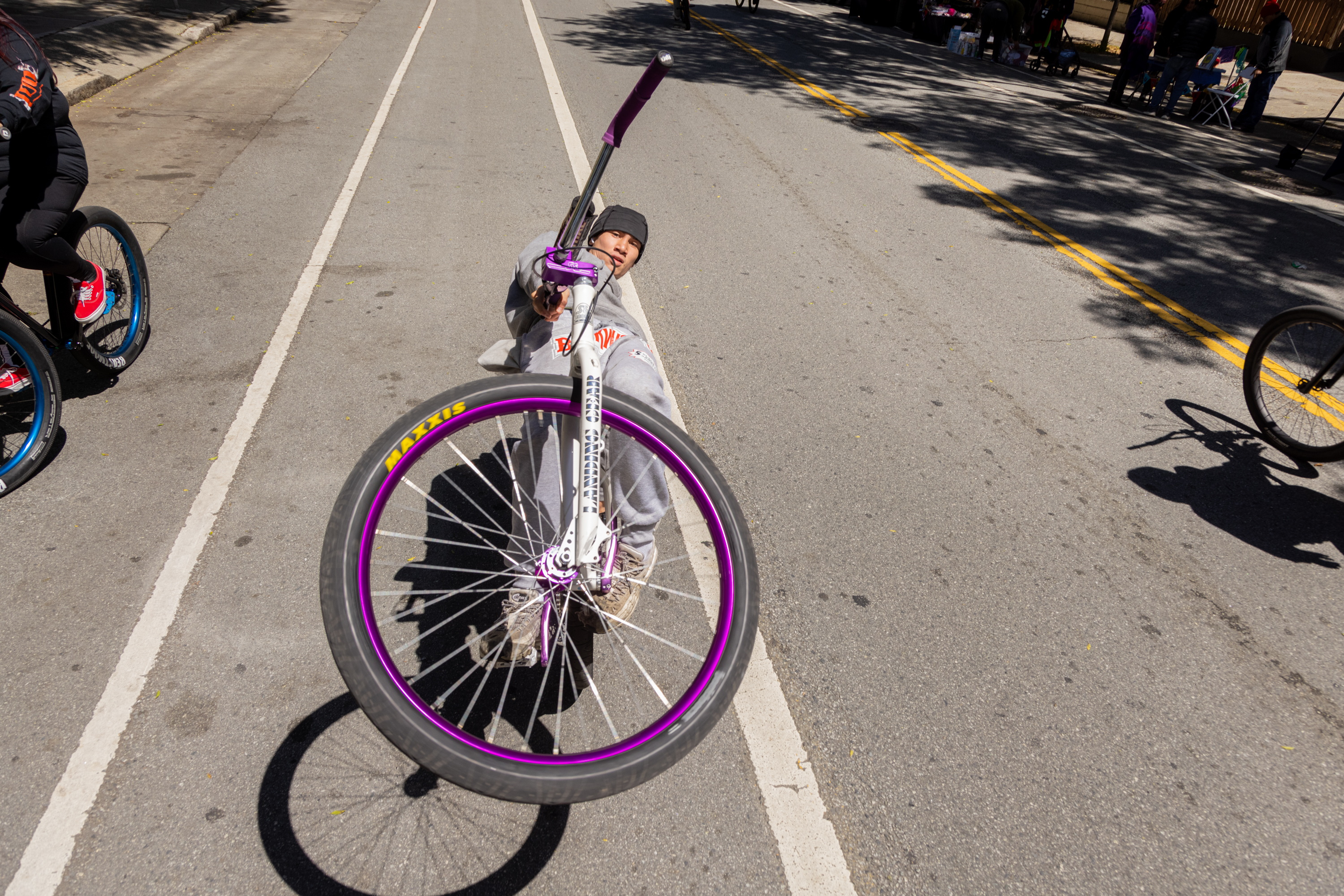 A child performs a bike trick, lifting the front wheel towards the camera, on a sunny street with onlookers.