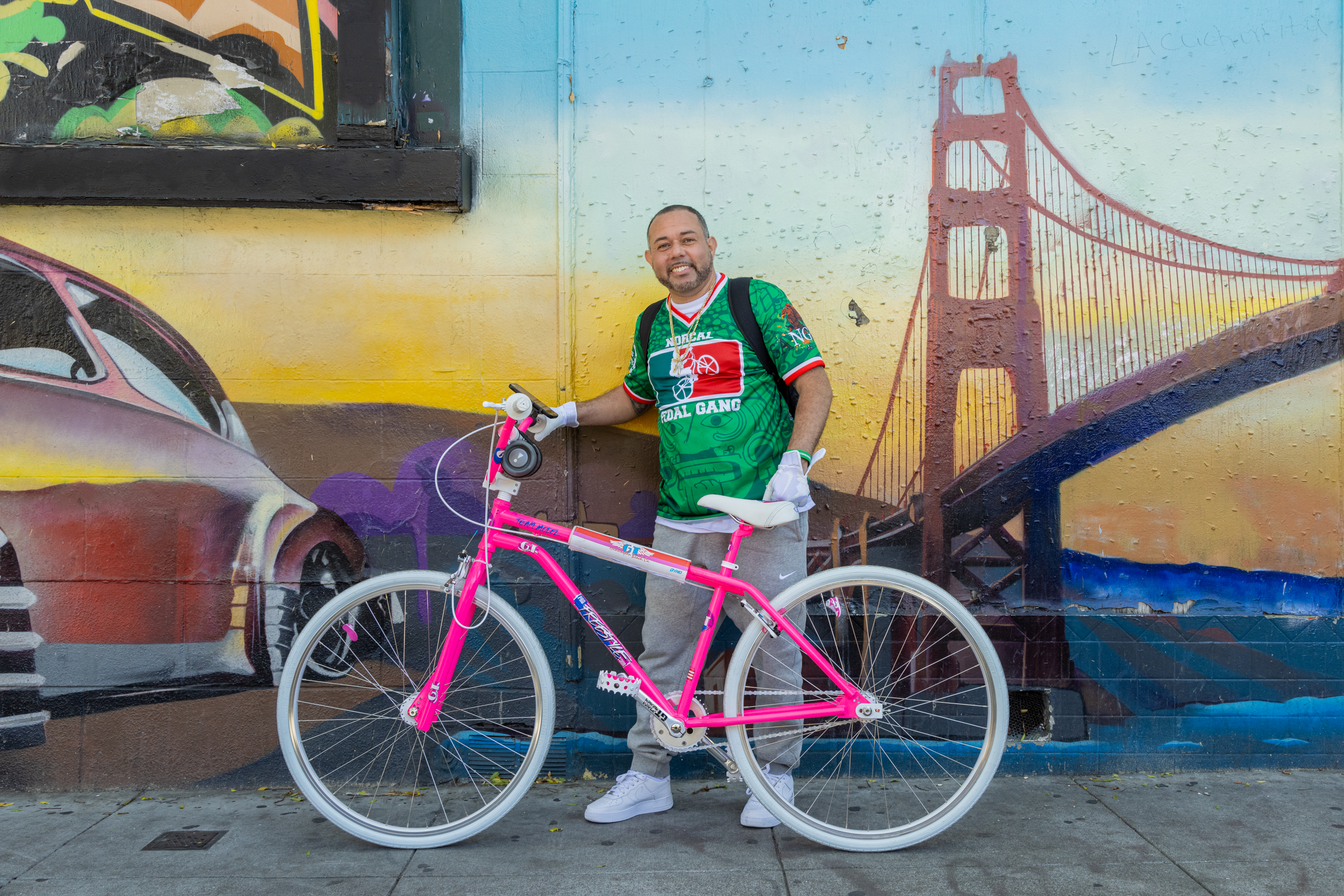 A man stands by a pink bike against a mural with a depiction of the Golden Gate Bridge.
