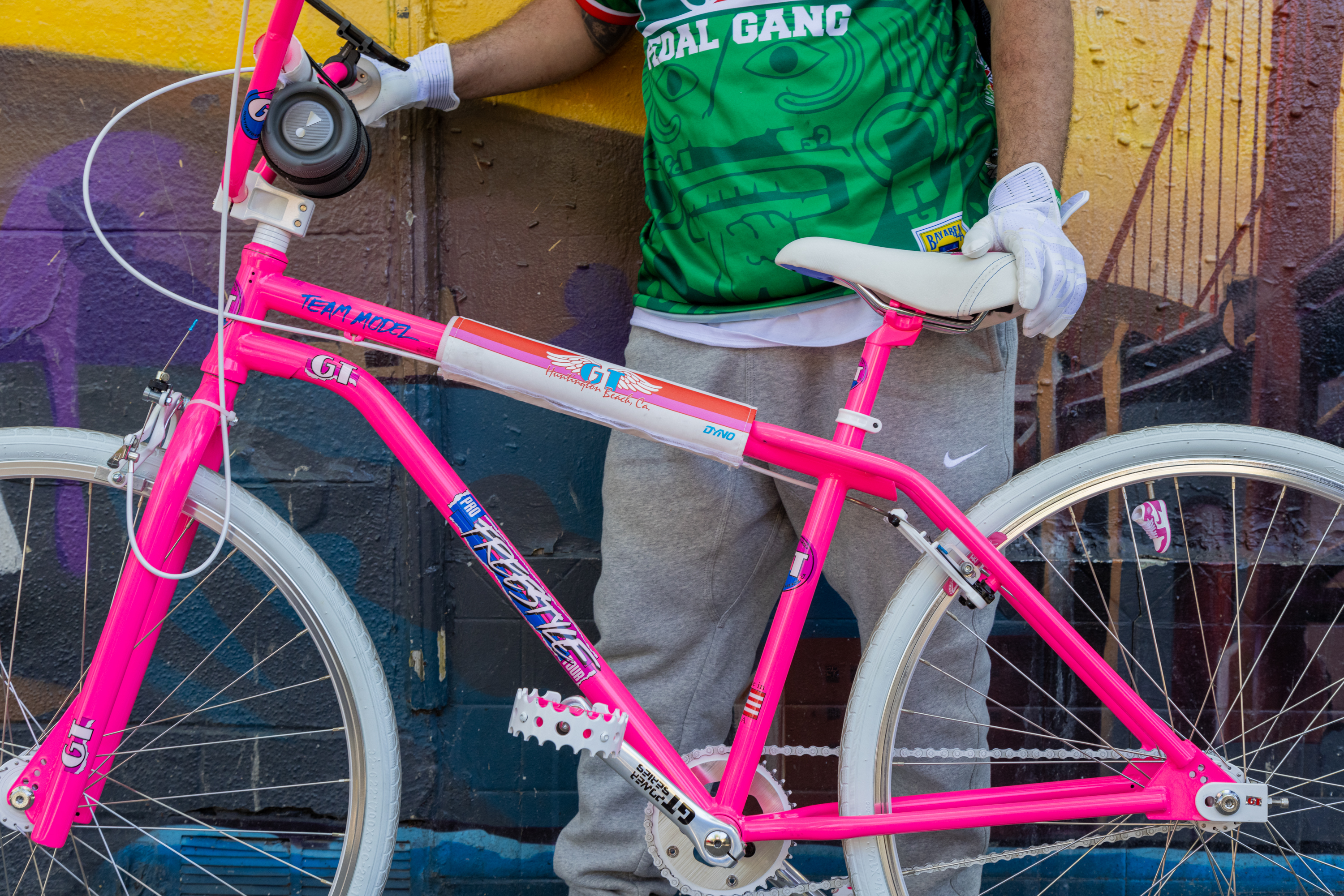 A person in a green jersey holds a pink bicycle against a colorful graffiti wall.