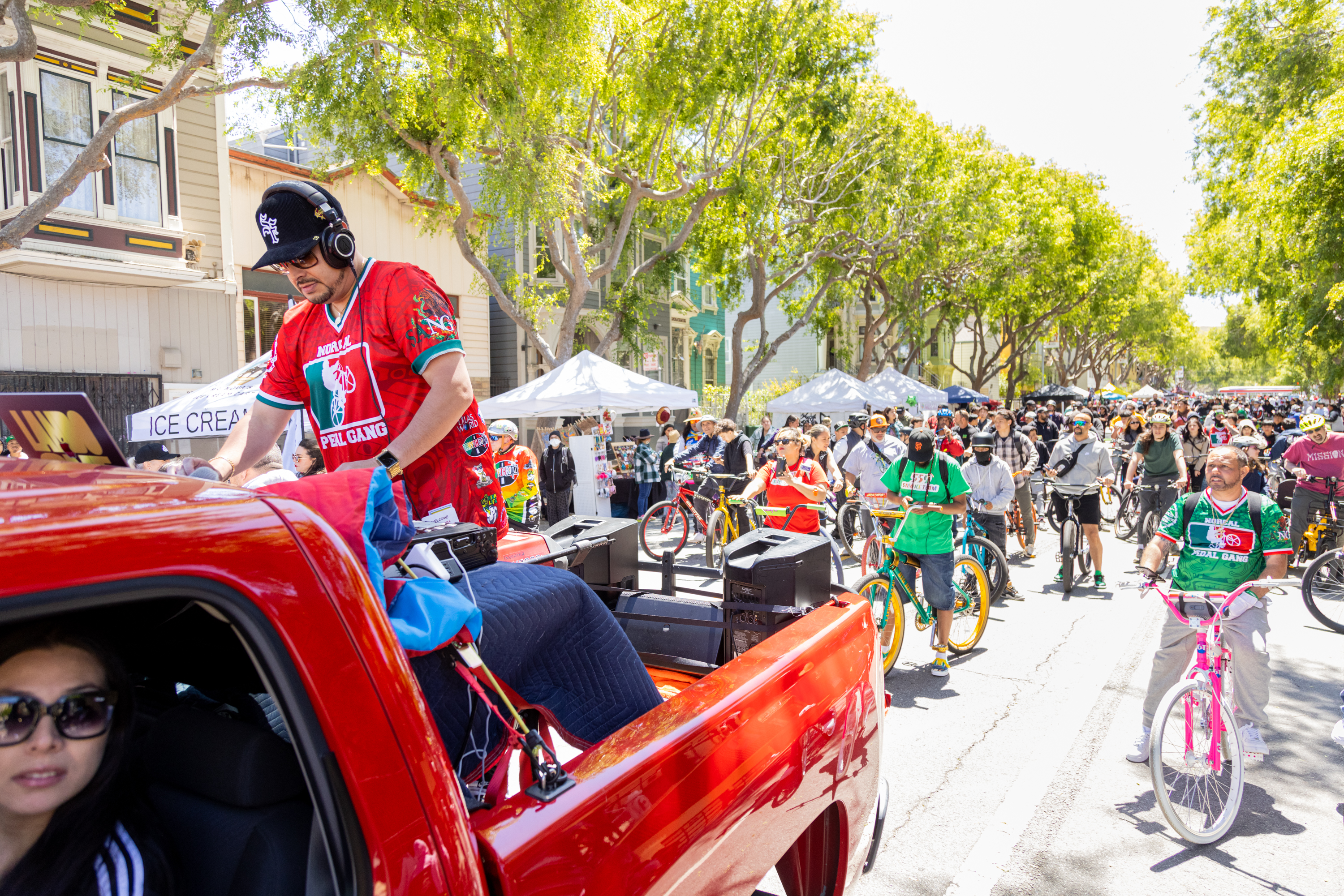 A DJ on a red truck plays music for a crowd of cyclists on a sunny street lined with trees and tents.