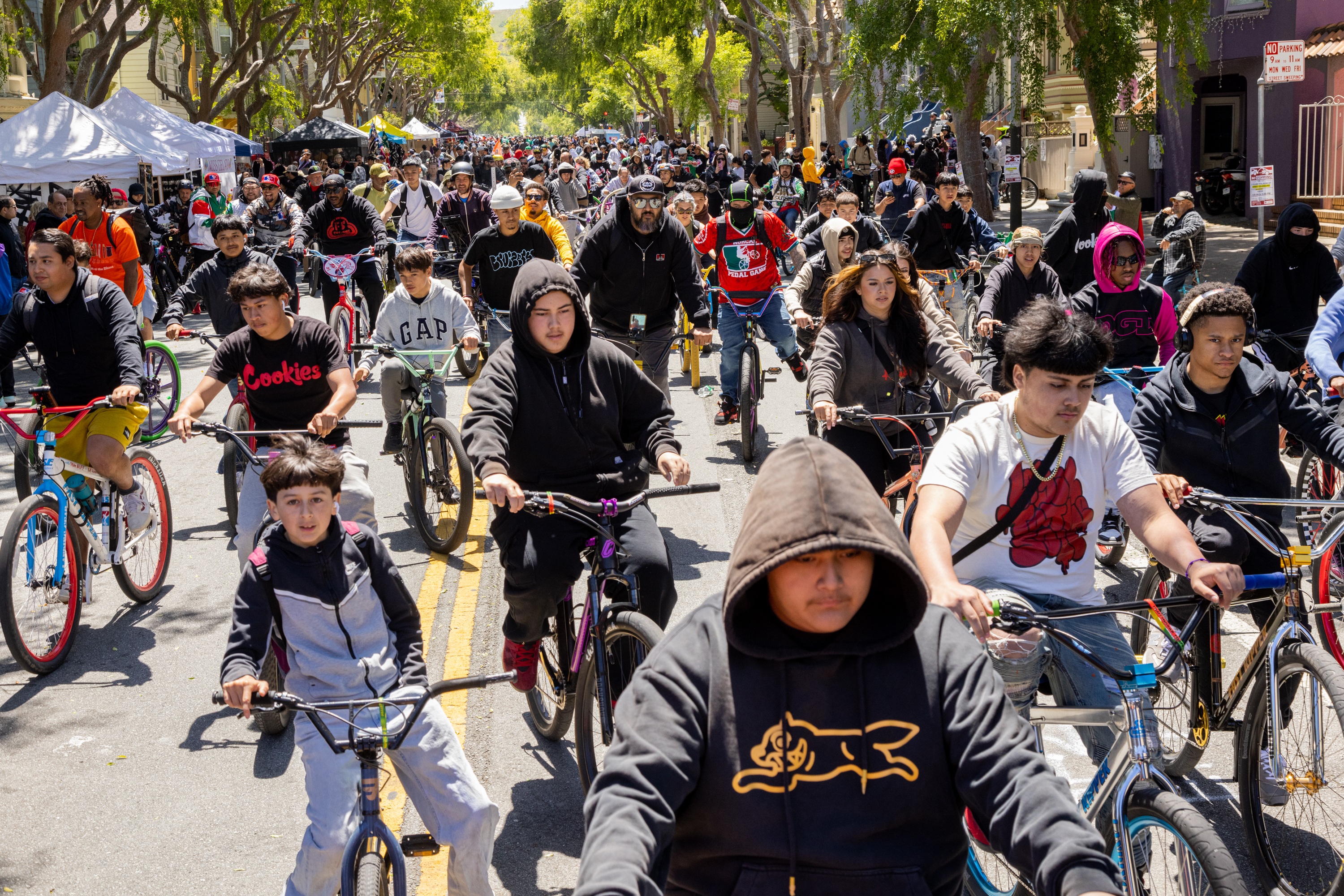 A diverse crowd cycles down a sunny, tree-lined street, creating a dynamic sea of riders and bicycles.