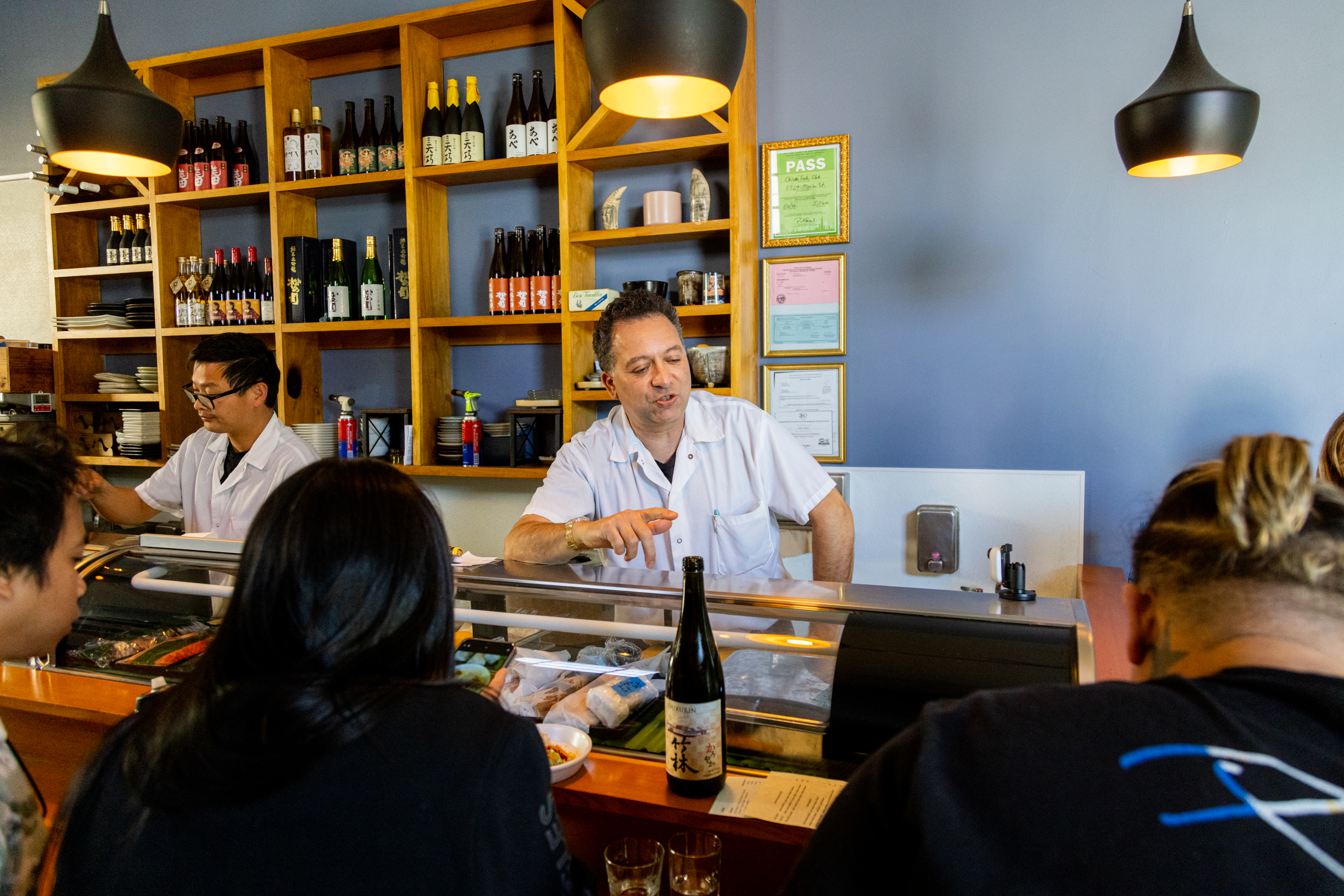 A chef in a white shirt talks to customers at a sushi bar. Bottles and dishes are displayed on shelves behind him. Another chef works nearby, preparing food.