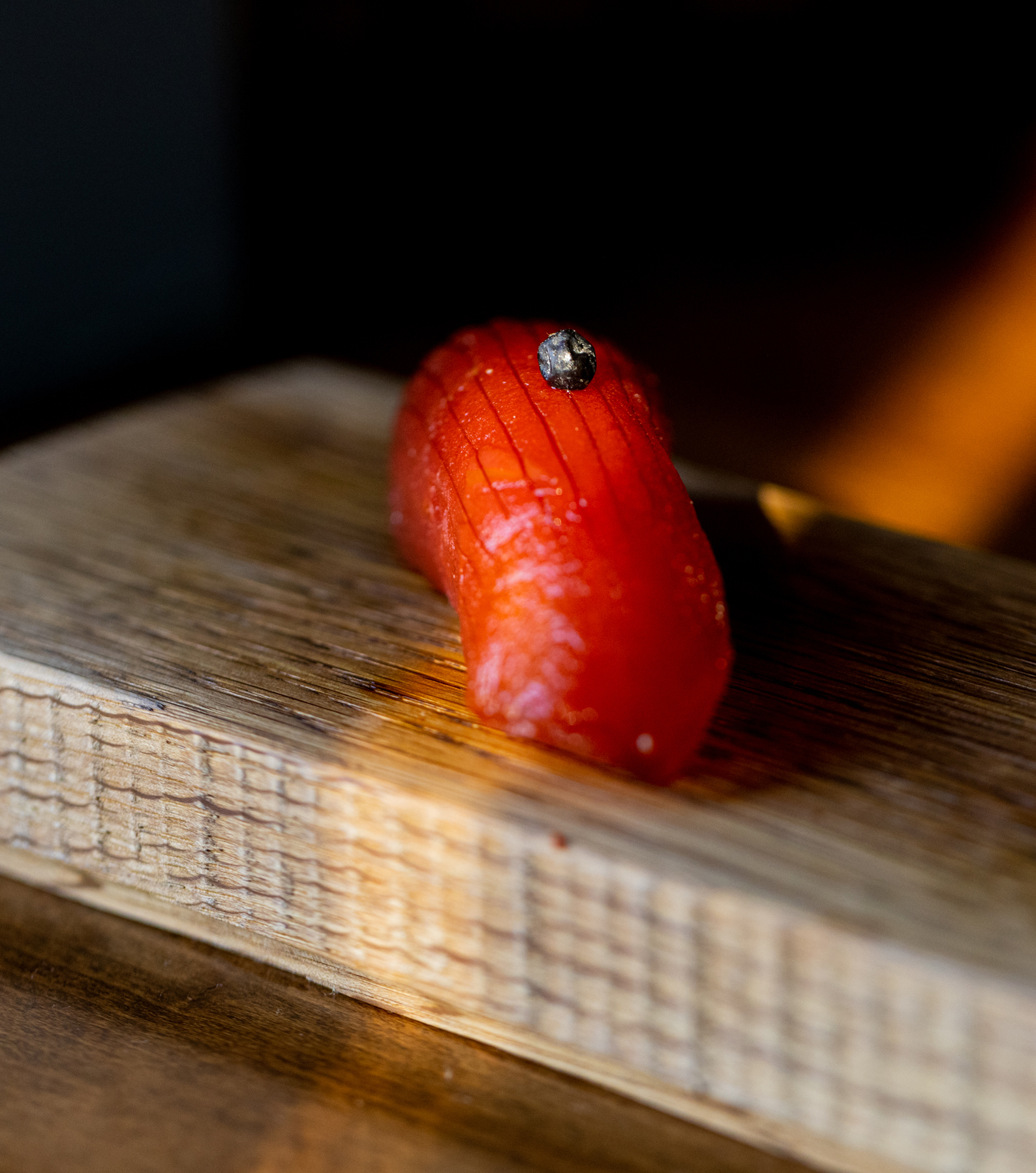 A piece of finely sliced red fish sushi, garnished with a small black element, is placed on a textured wooden block, illuminated warmly.