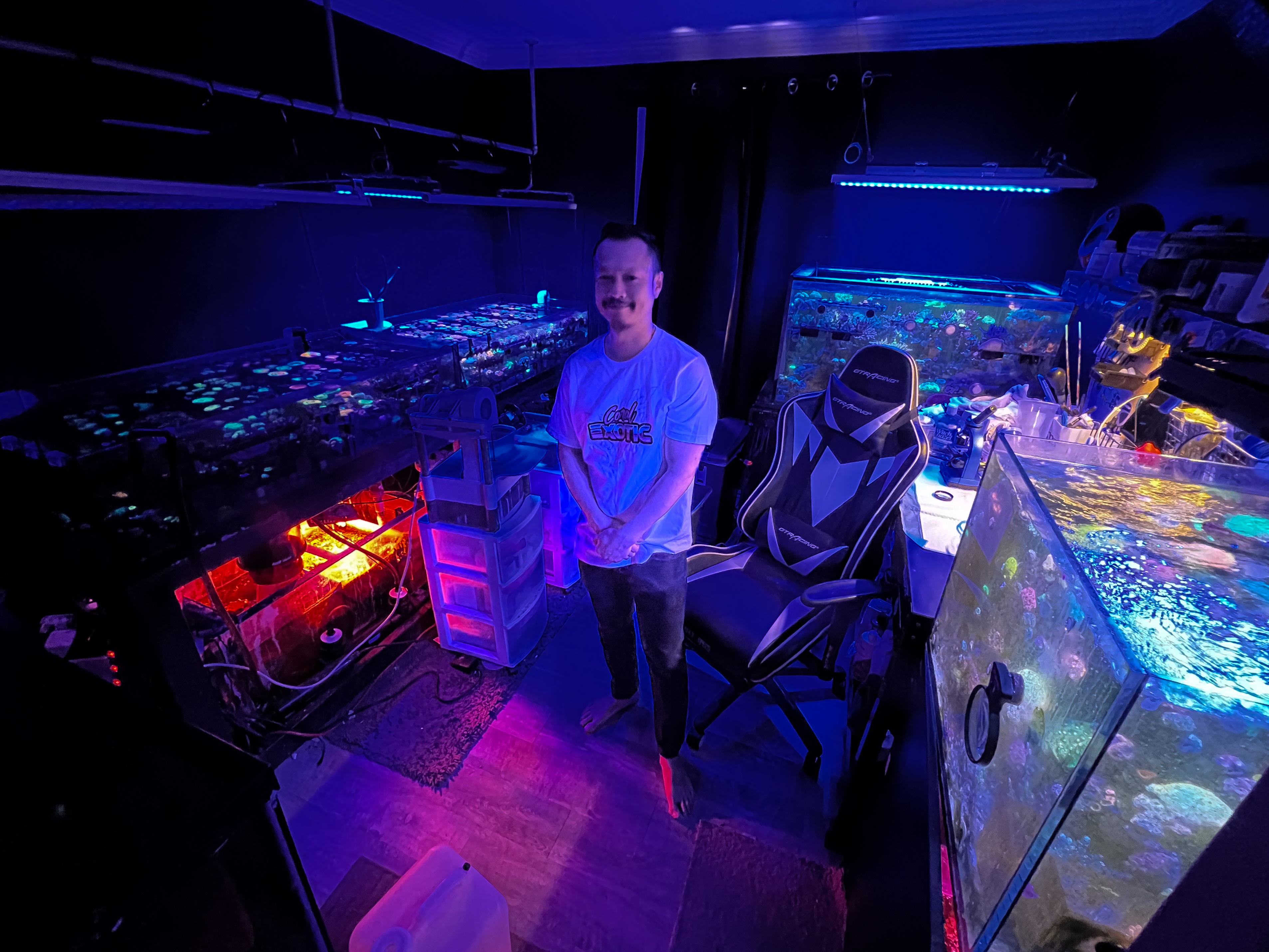 A man stands in a room lit by blue lights, surrounded by vibrant aquariums and coral tanks, with a gaming chair to the right.