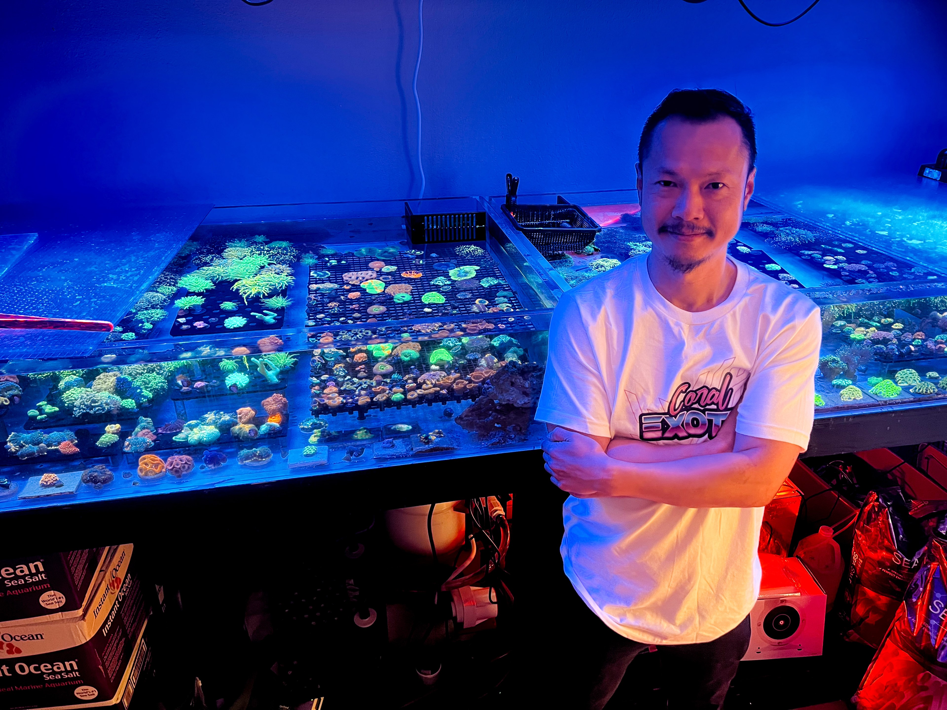 A man smiles, standing beside vibrant coral tanks under blue lighting, arms crossed, wearing a white T-shirt.