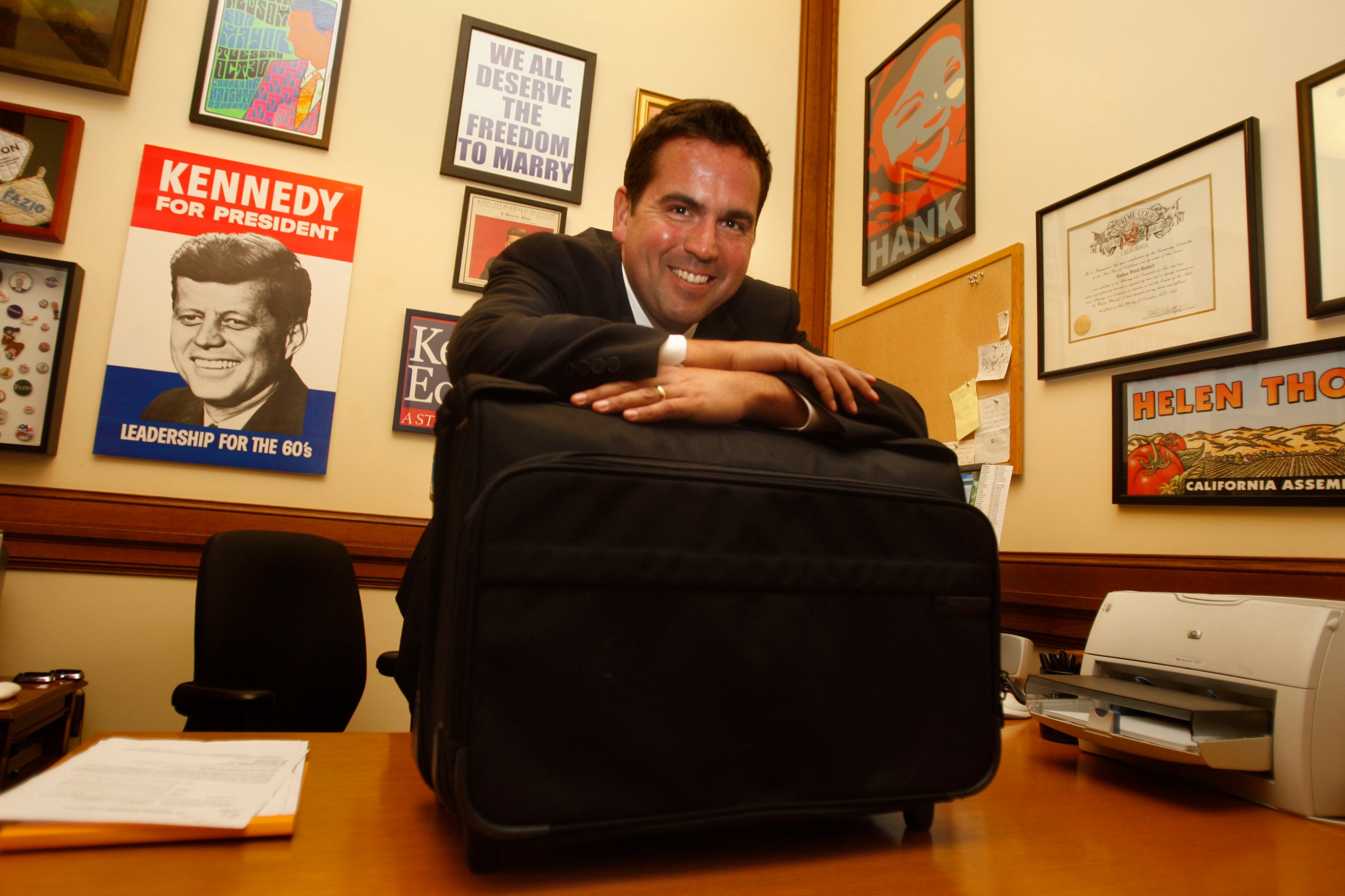 Nathan Ballard smiles as he wears a suit and leans on a briefcase in an office adorned with various political posters and certificates.