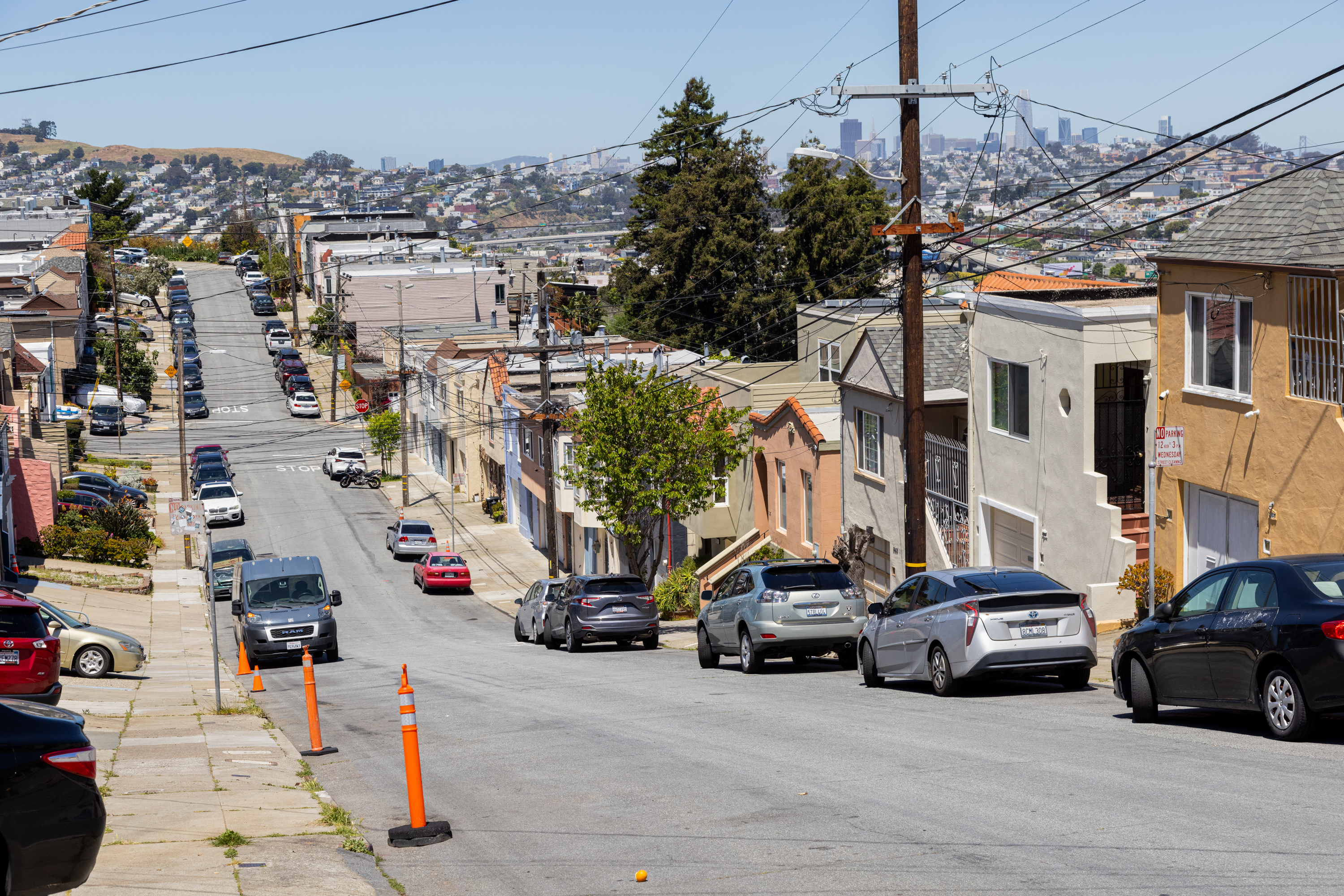 A steep street lined with parked cars and houses, with a city skyline in the distance.